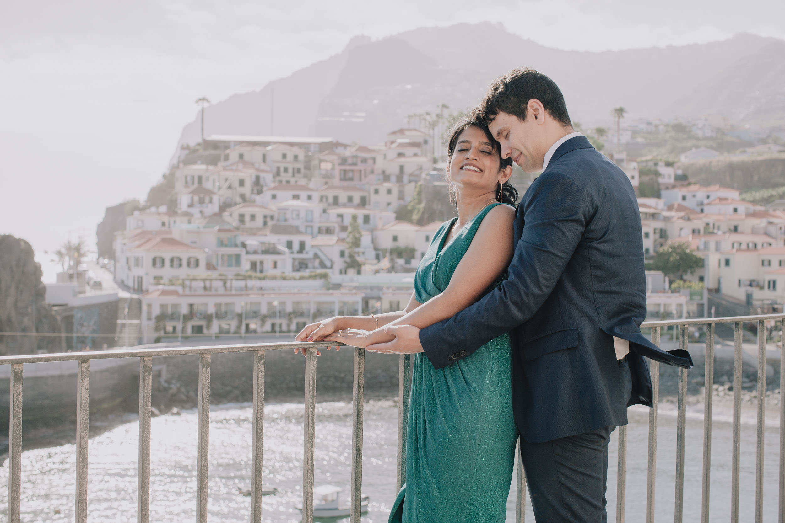 He holds her from behind at the railing, her eyes closed and her head against him, the village on the cliff behind, Camara de Lobos, Madeira
