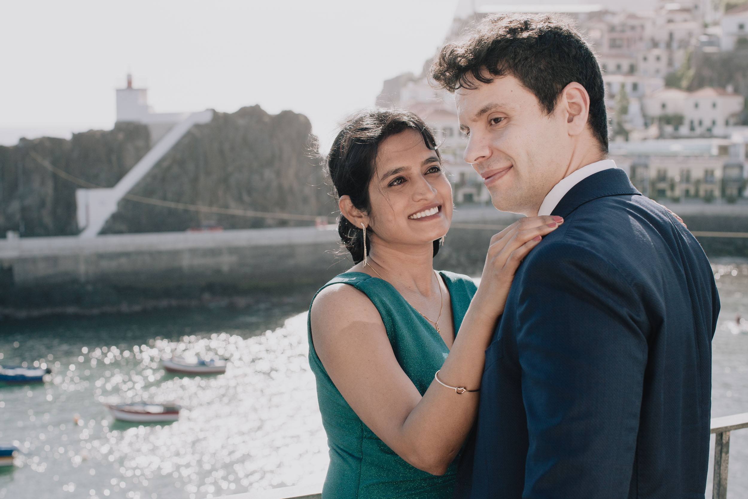 She looks up at him with her hand on his shoulder, the breakwater and the lighthouse out of focus behind, Camara de Lobos, Madeira