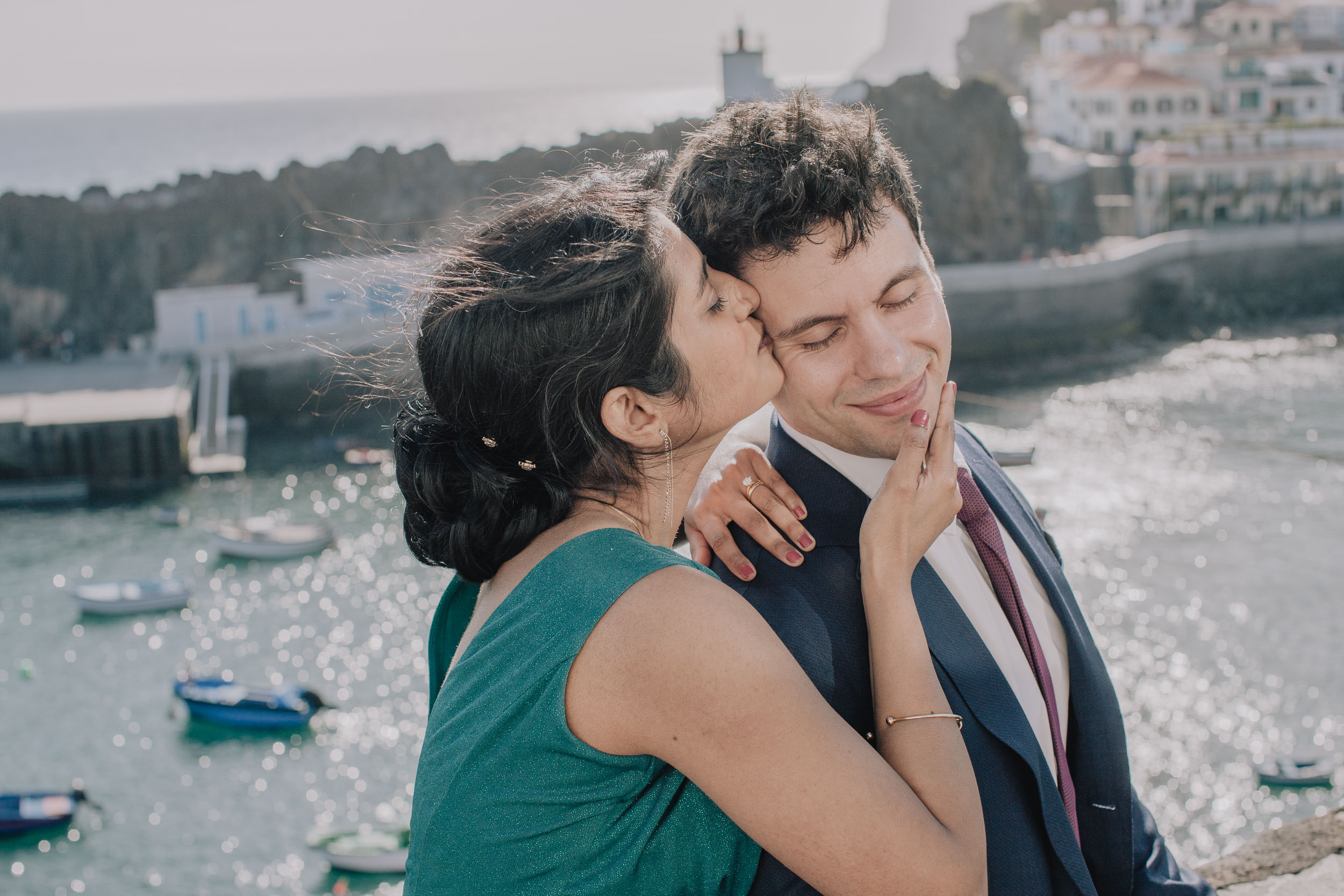 Bride kissing the groom on the cheek as he smiles, the harbour soft behind, Câmara de Lobos, Madeira