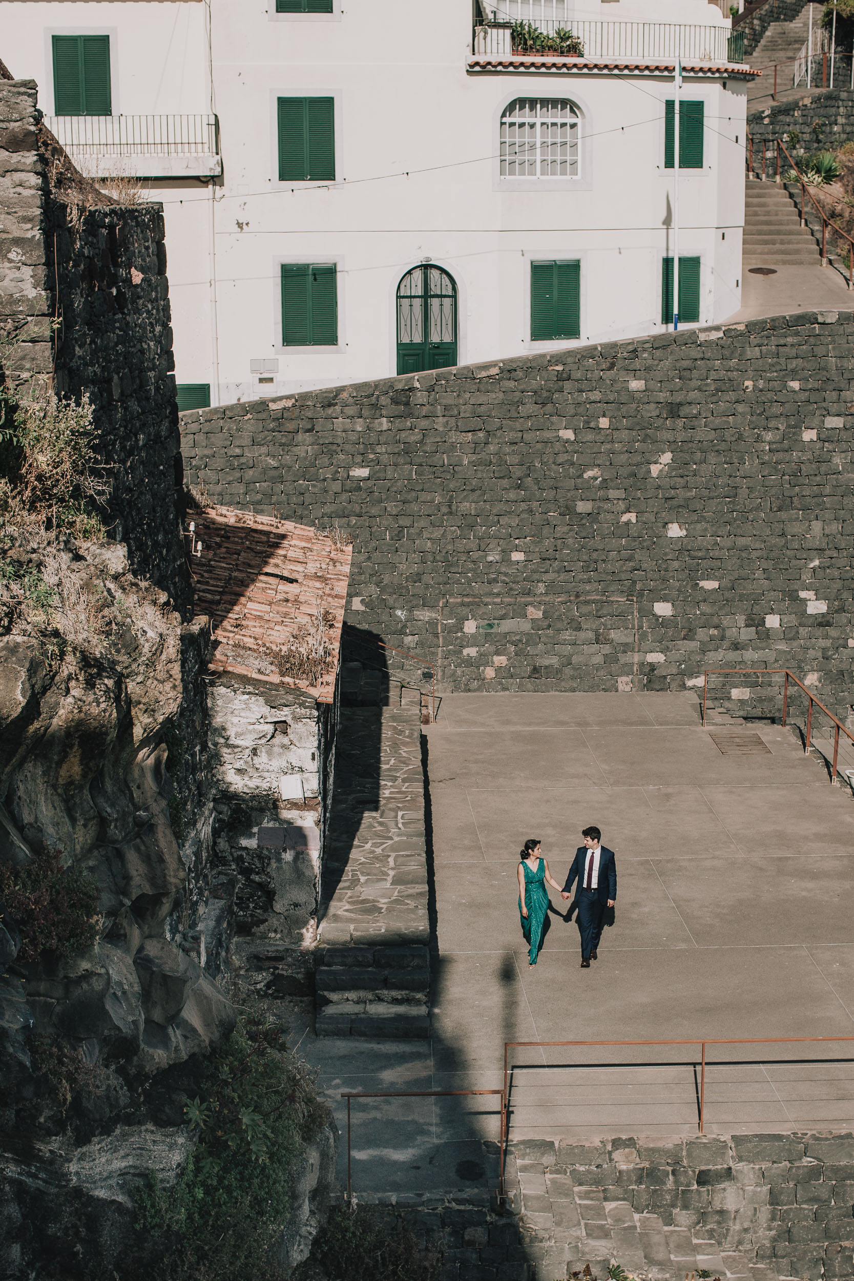 Couple walking hand in hand across a terrace below a white house with green shutters, Câmara de Lobos, Madeira