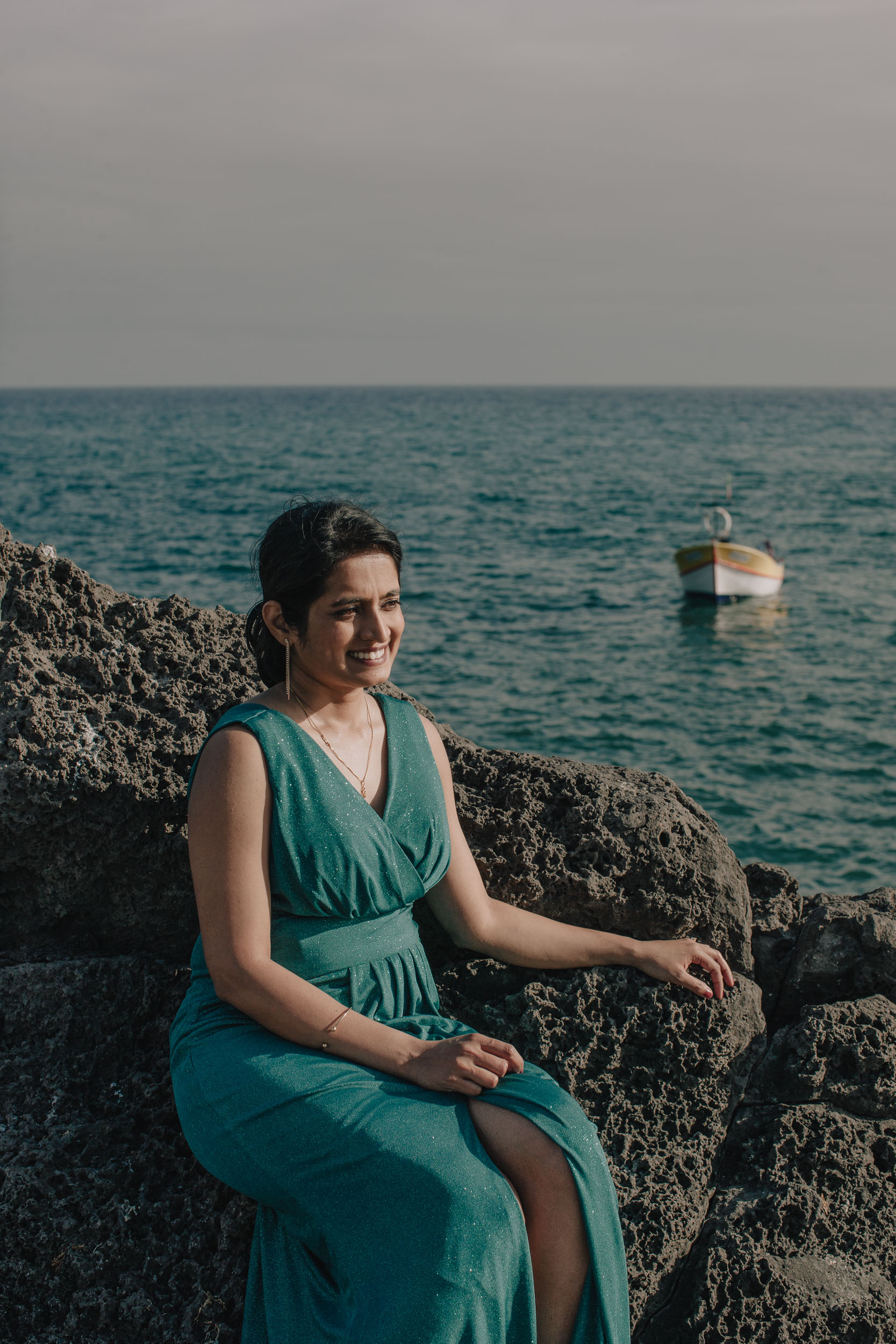 Bride in a green dress sitting on the volcanic rocks by the sea, a lone fishing boat behind, Câmara de Lobos, Madeira
