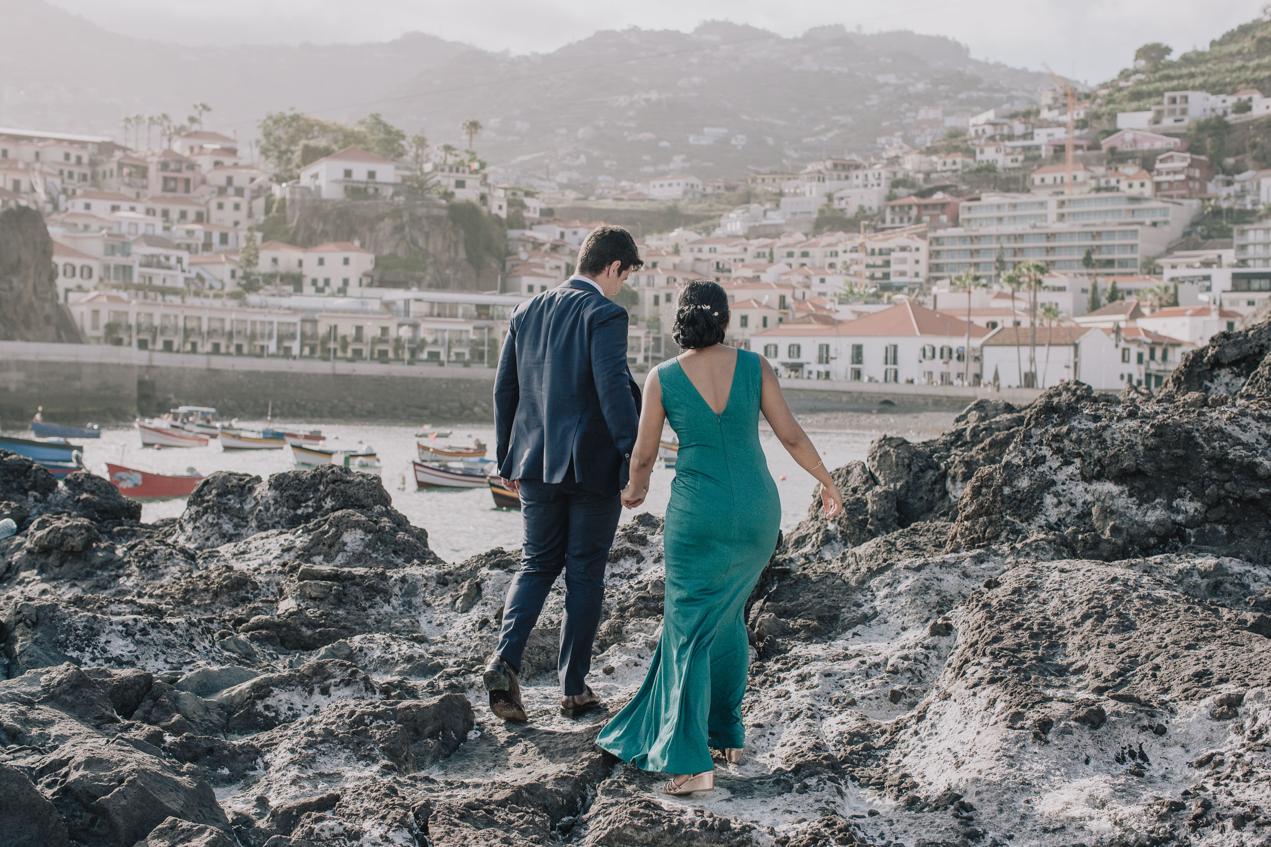 Couple walking hand in hand across the volcanic rocks toward Câmara de Lobos village, seen from behind, Madeira