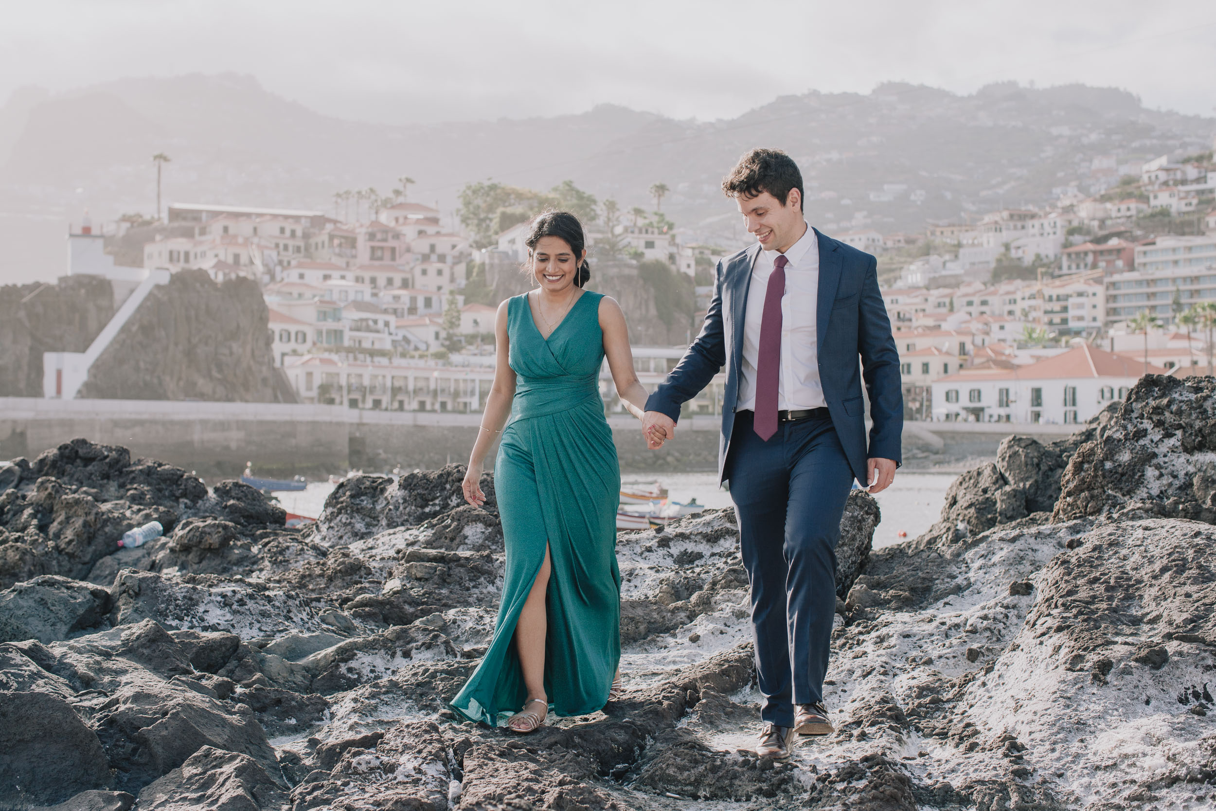 Couple walking hand in hand across the rocks, both smiling, Câmara de Lobos village and mountains behind, Madeira