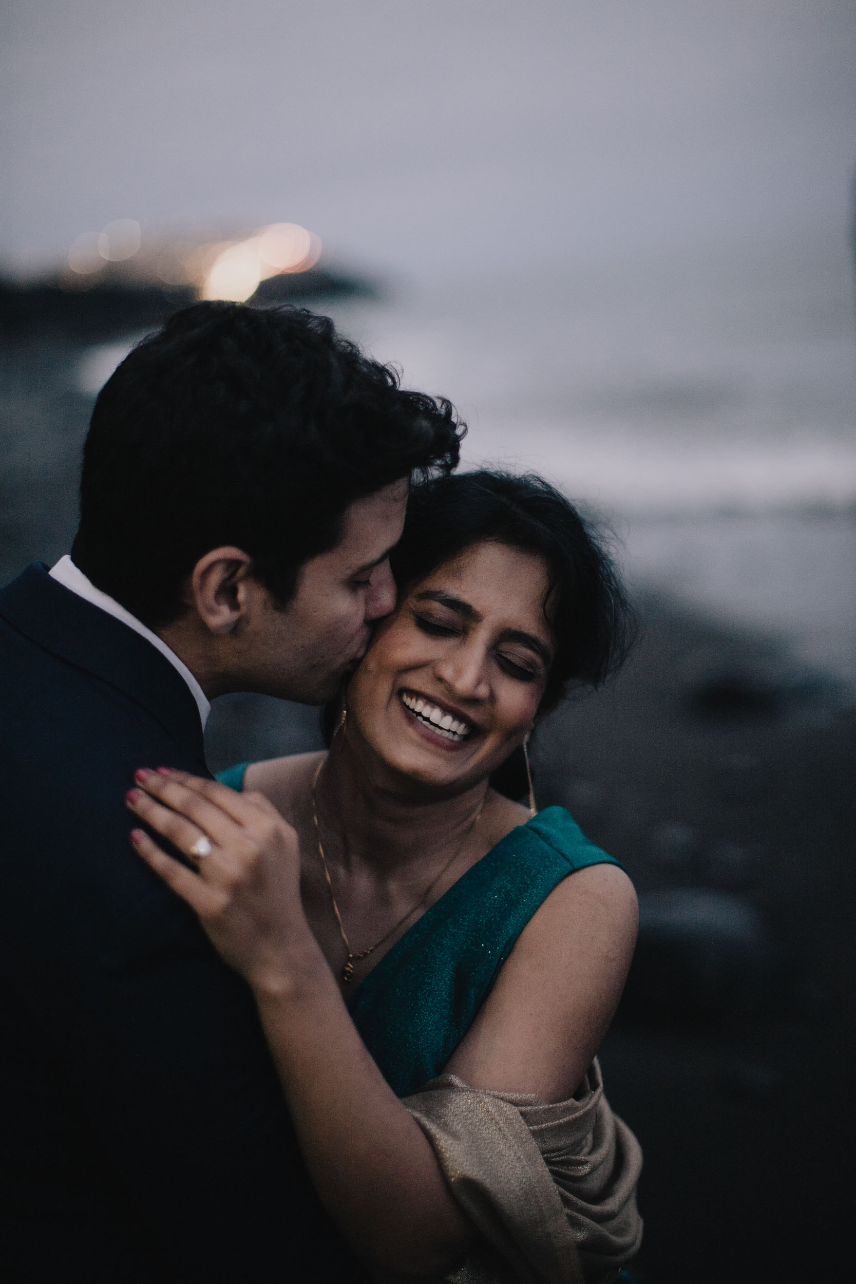 Groom kissing the bride's cheek at dusk as she laughs, the lights of the pier behind, Câmara de Lobos, Madeira