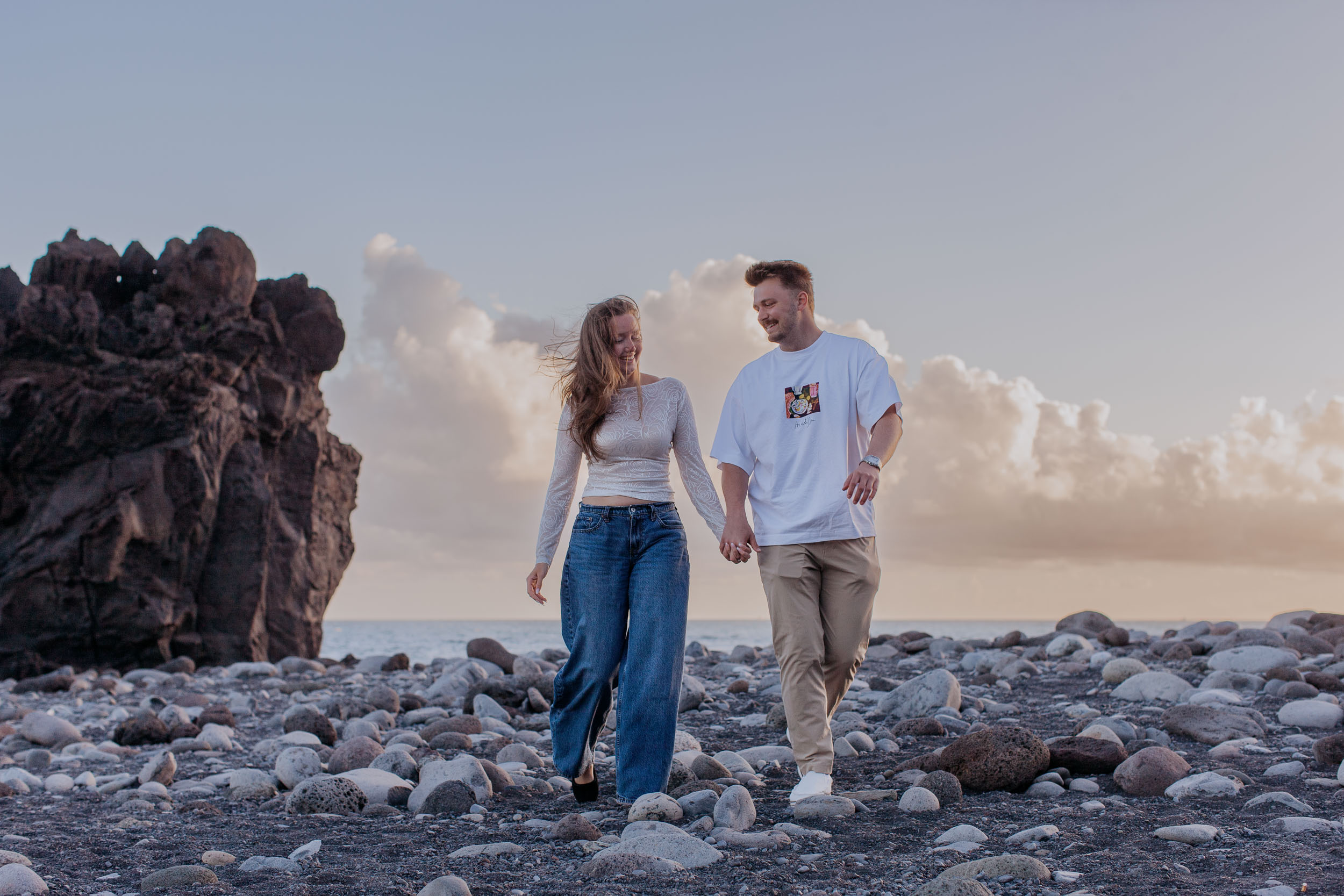 Rylie and Abby walking hand in hand on the round black pebbles of Praia Formosa, a tall volcanic outcrop and pastel sunset clouds behind, Madeira