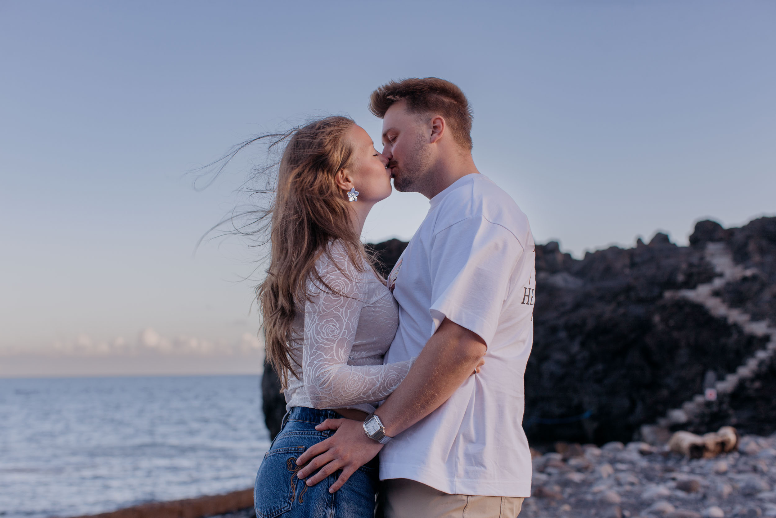 Kissing in profile on the pebble beach, her hand at his waist, wind lifting her hair, Praia Formosa, Madeira