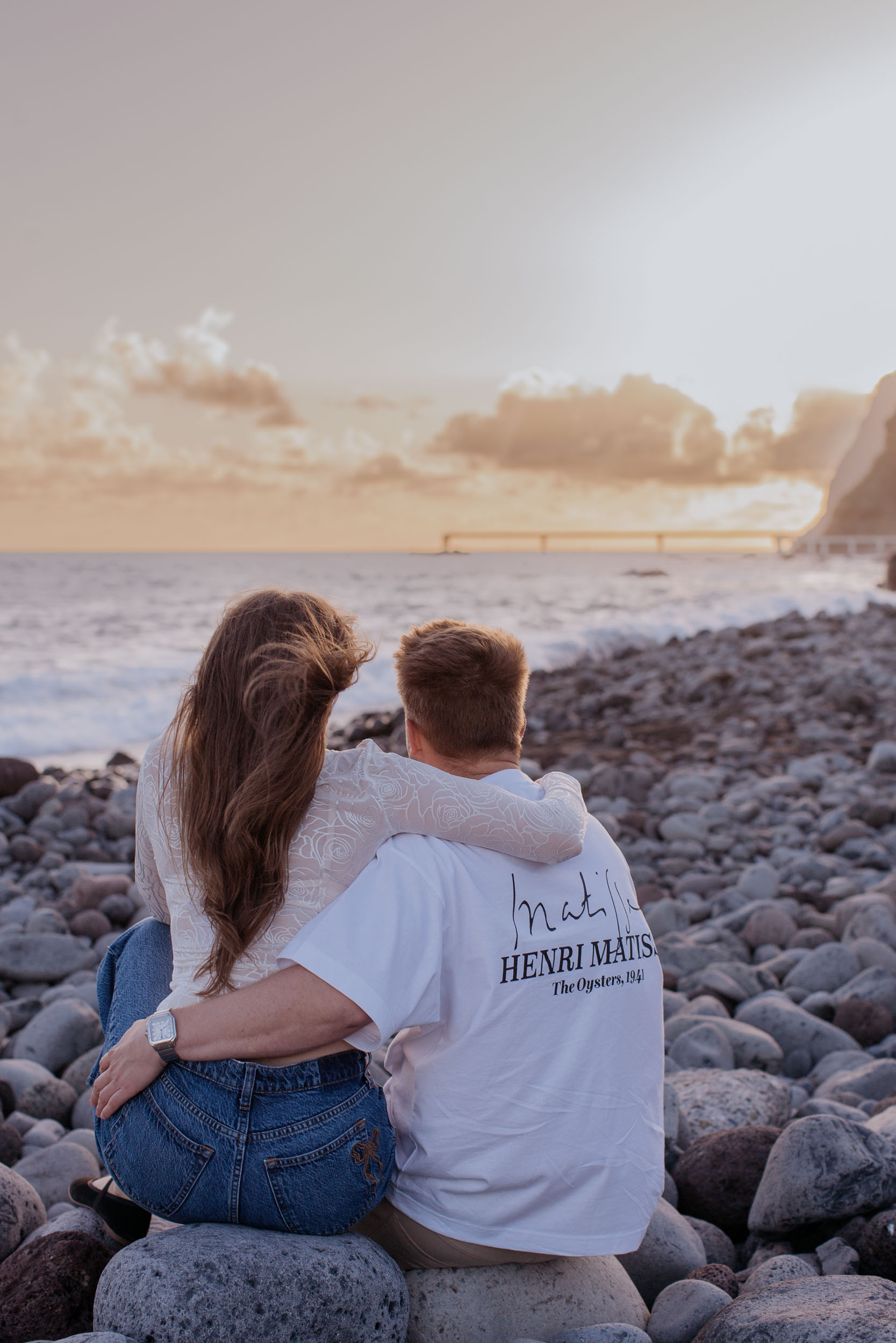Rylie and Abby sitting on the pebble shore with his arm around her, watching the sun fall toward the Atlantic at Praia Formosa, Funchal, Madeira