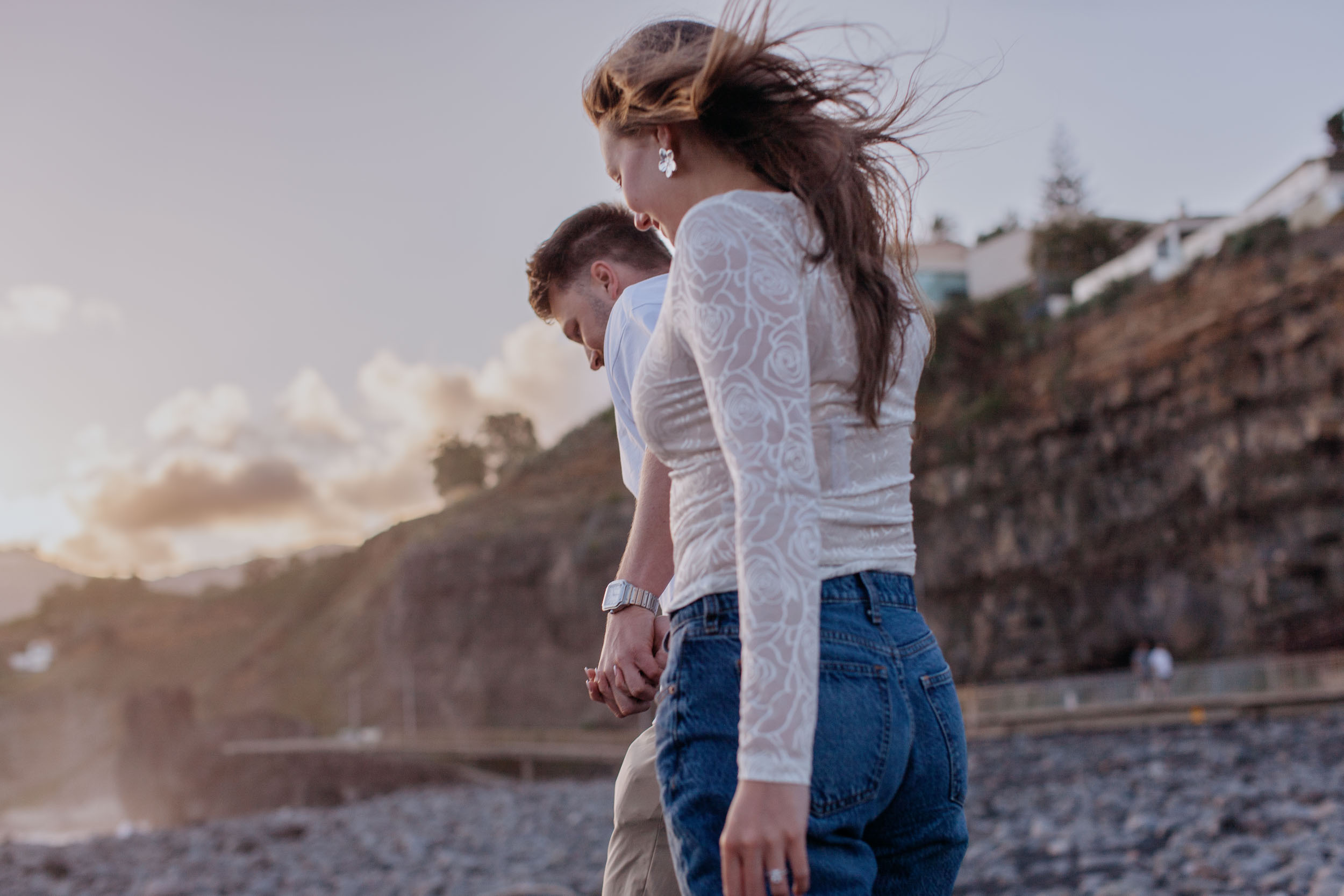 Close from behind as she walks past the camera in a white lace top, he follows with his head down, hands joined, Praia Formosa, Madeira