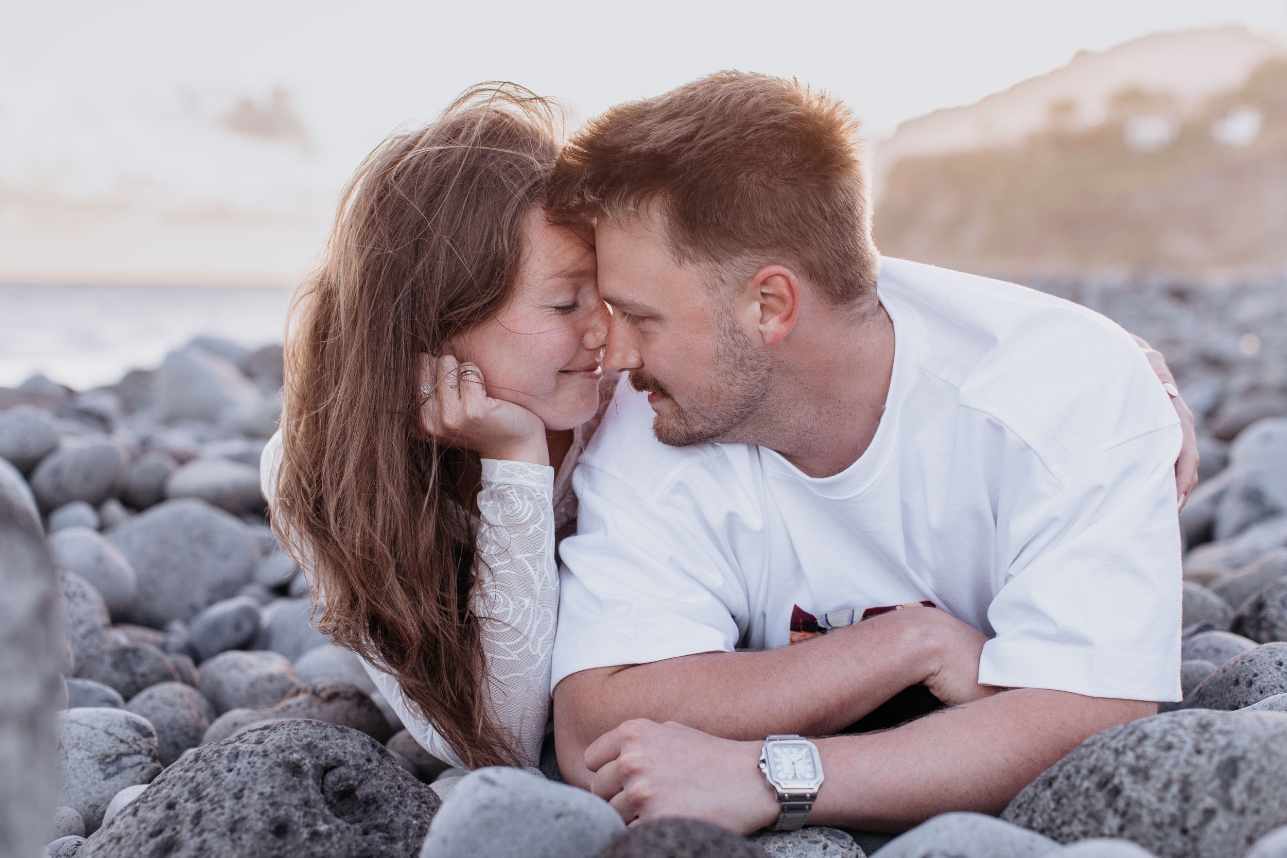 Noses almost touching as they lie on the pebbles, both smiling with eyes shut, Praia Formosa, Madeira