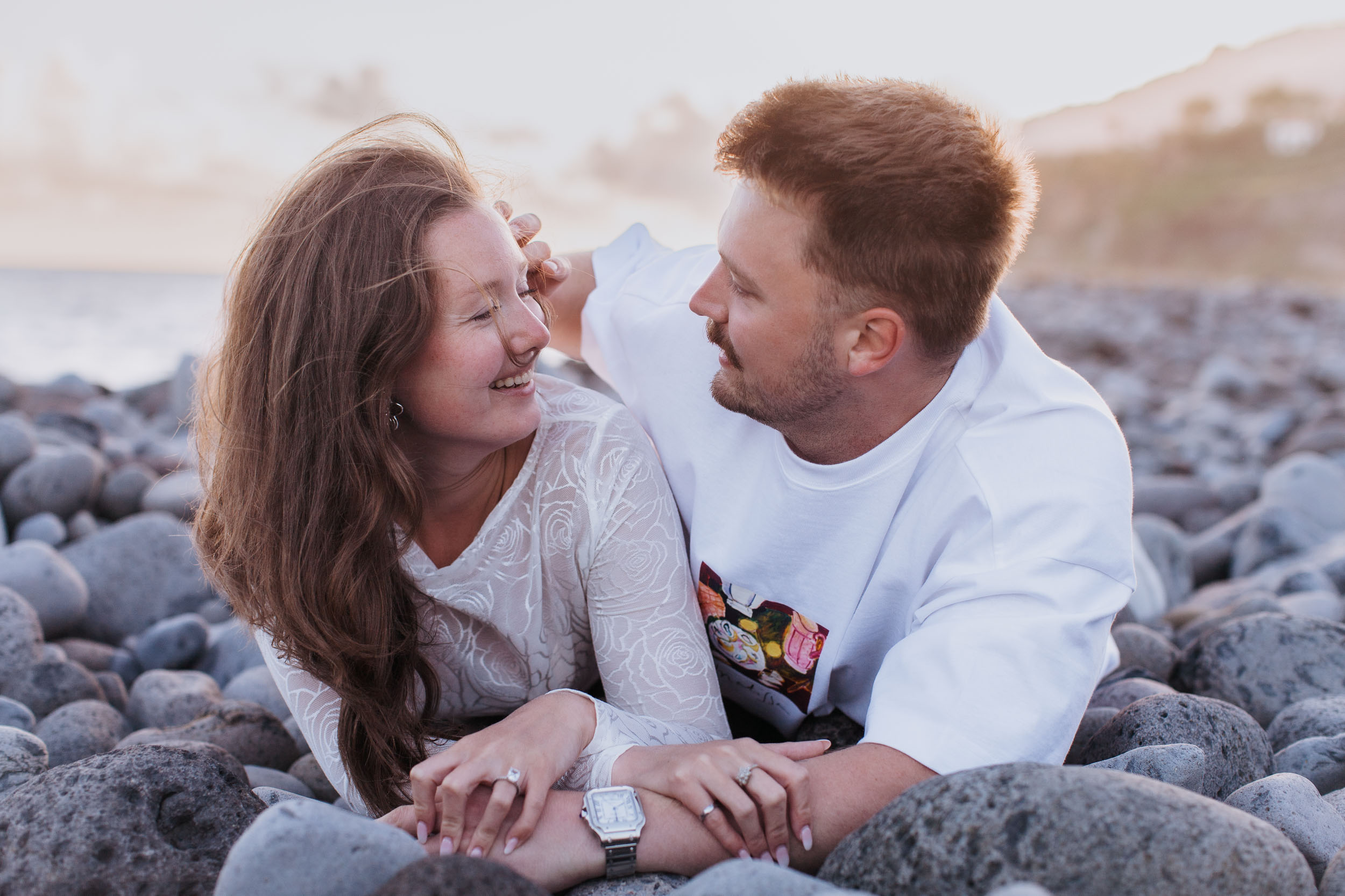 She laughs with her eyes closed while he brushes the hair off her face, both lying on the stones, Praia Formosa, Madeira