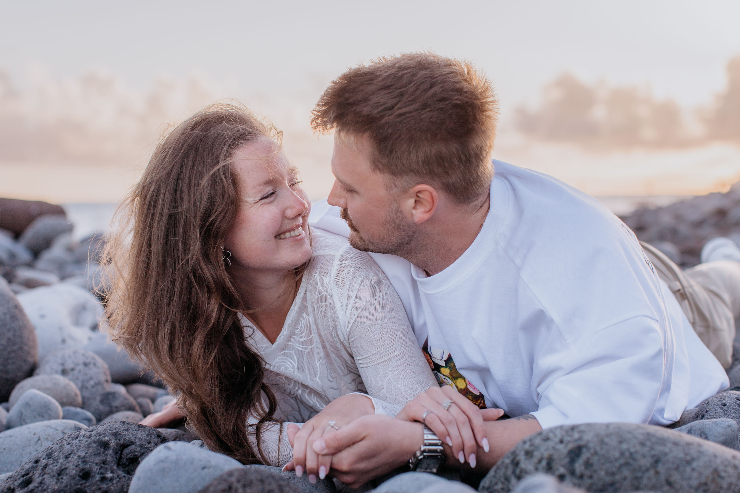 Abby laughing on Rylie's chest as they lie on the black pebbles, warm late light across their faces, Praia Formosa, Madeira