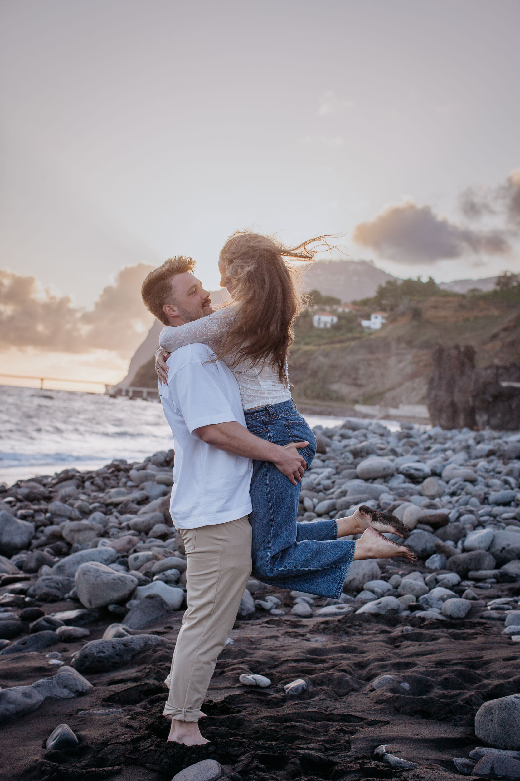 Vertical frame. He lifts her off the ground in an embrace, her feet swinging, the cliff and the lit village behind, Praia Formosa, Madeira