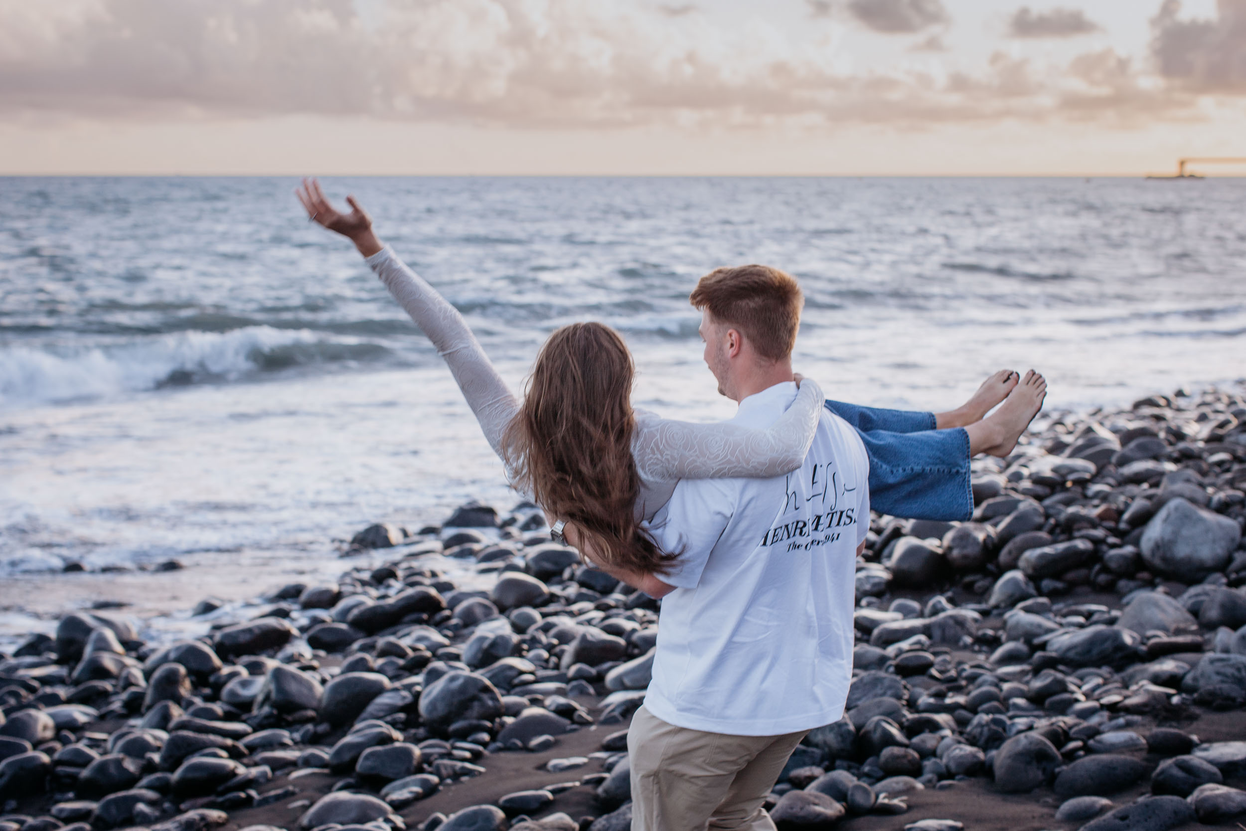 Seen from behind, he carries her along the stones and she throws one arm wide toward the surf, Praia Formosa, Madeira