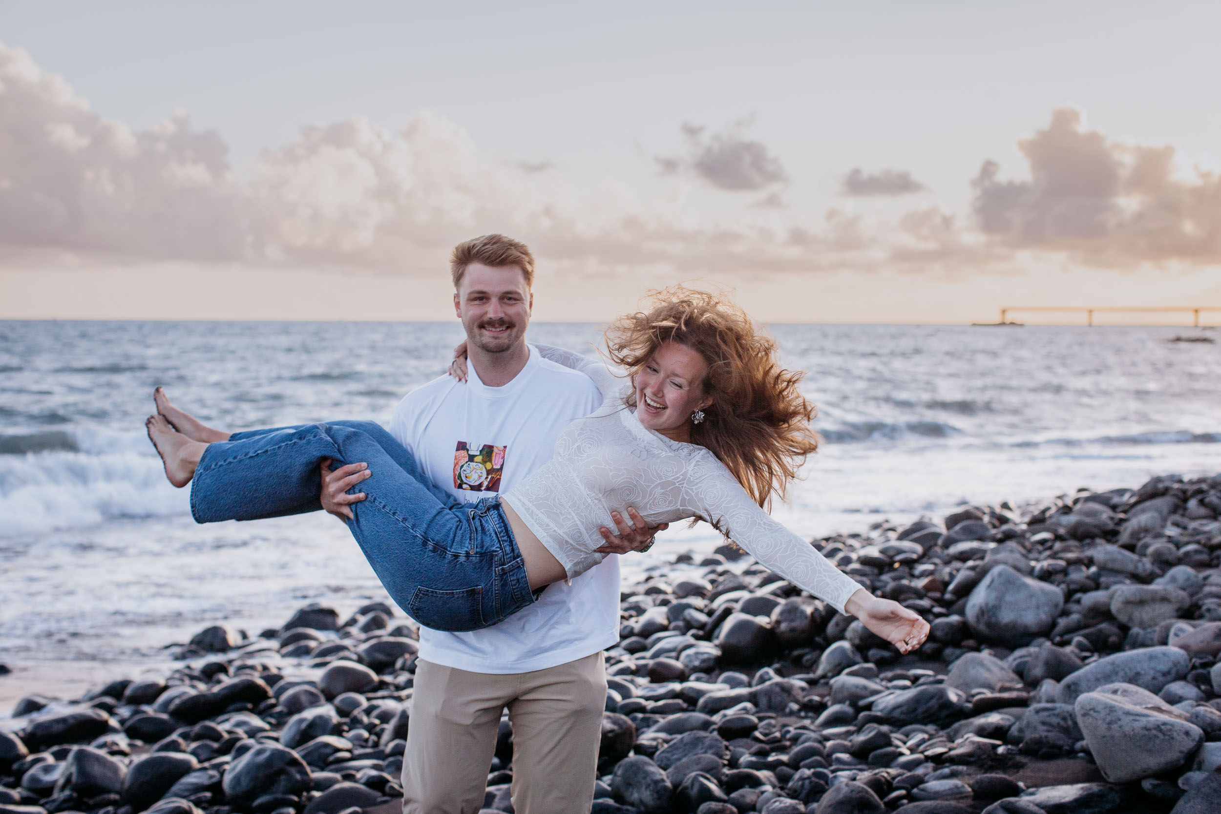 Rylie carrying Abby in his arms across the pebble beach, both laughing into the wind, sunset clouds and Funchal pier behind, Praia Formosa, Madeira