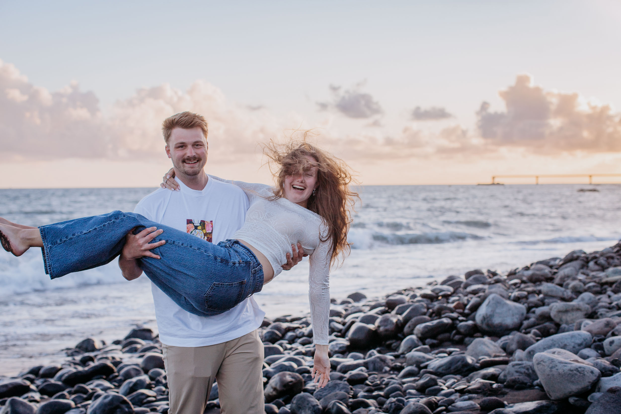 He carries her across the pebbles and she lets her head fall back laughing, hair hanging down, Praia Formosa, Madeira
