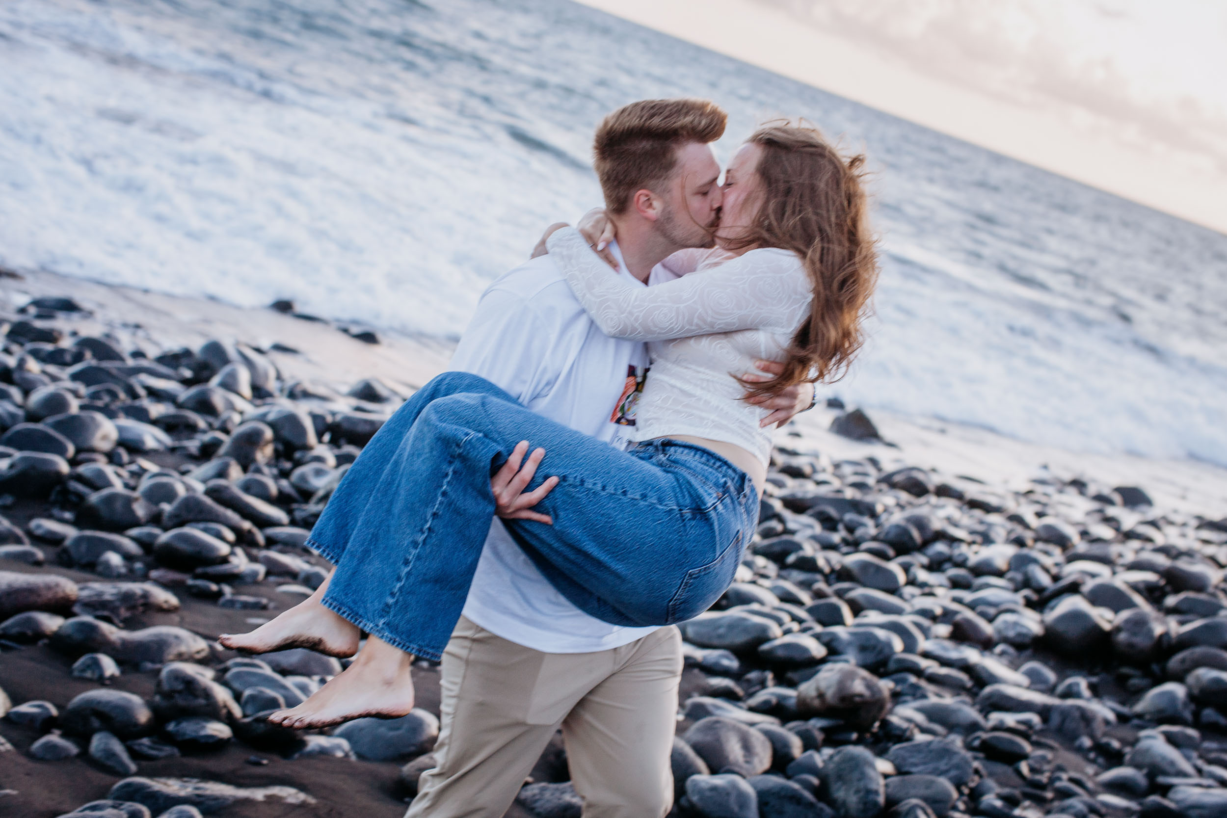 Rylie lifting Abby off the ground as they kiss on the pebble shoreline, ocean and stones behind them, Praia Formosa, Madeira
