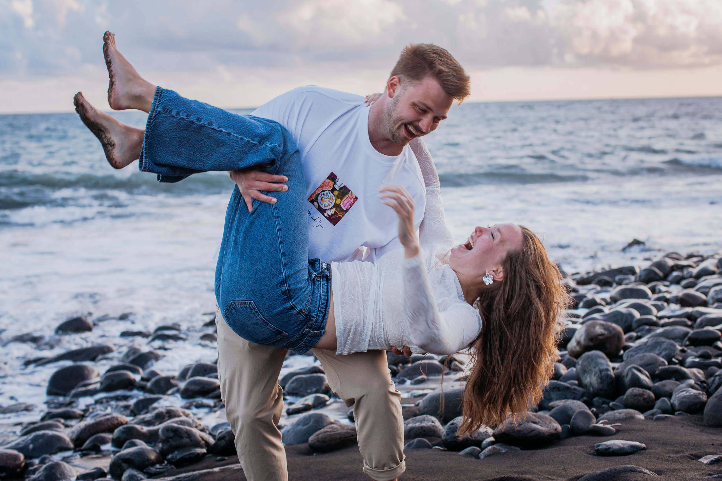 He holds her sideways in his arms and she tips her head back mid-laugh, bare feet in the air, Praia Formosa, Madeira