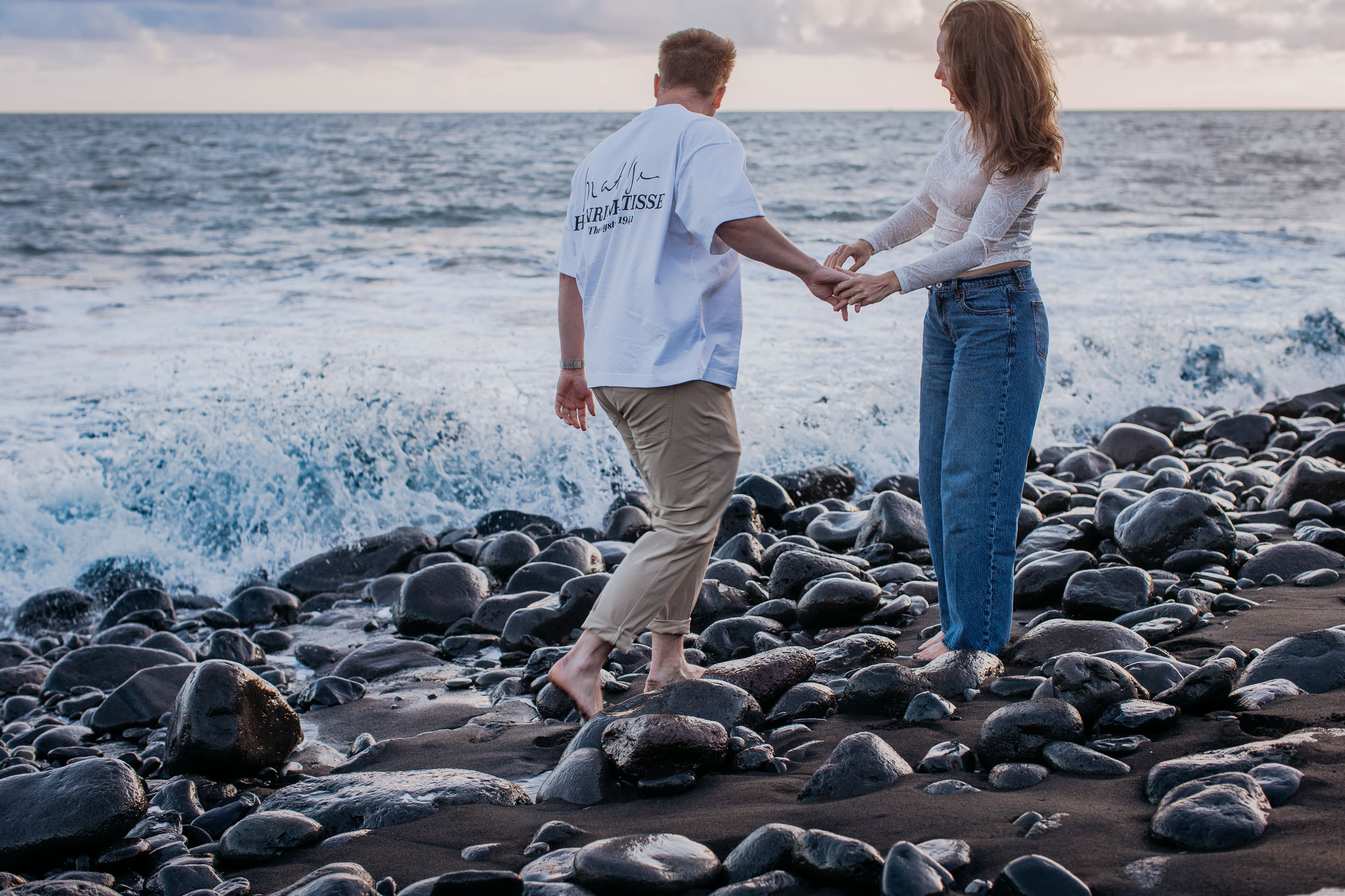 Rylie and Abby stepping carefully across wet volcanic stones as the surf rolls in beside them, Praia Formosa, Funchal, Madeira