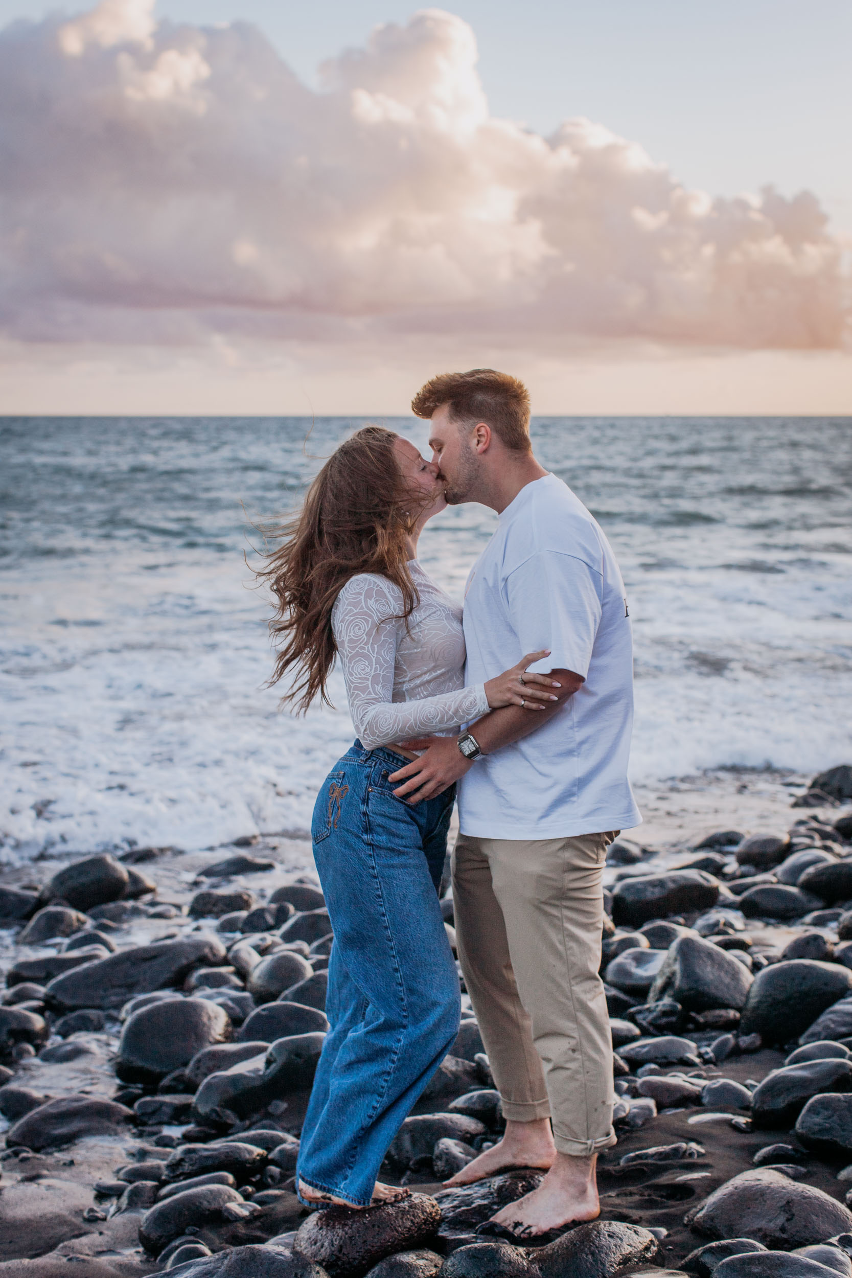 Vertical frame. A kiss at the waterline, the surf white behind them and pink cloud above the horizon, Praia Formosa, Madeira