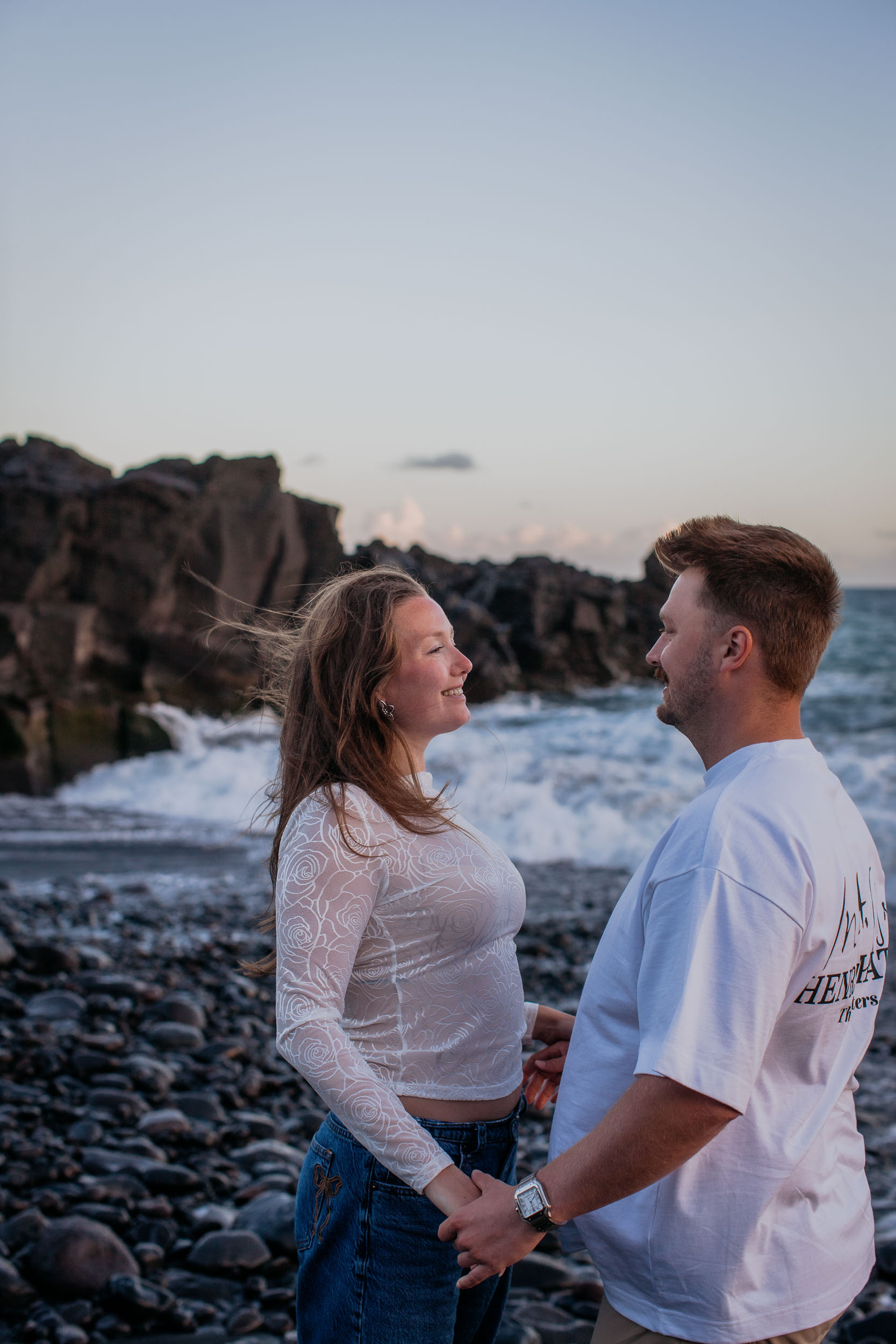 Abby smiling up at Rylie as they hold hands by the volcanic cliff and breaking surf, golden hour at Praia Formosa, Madeira