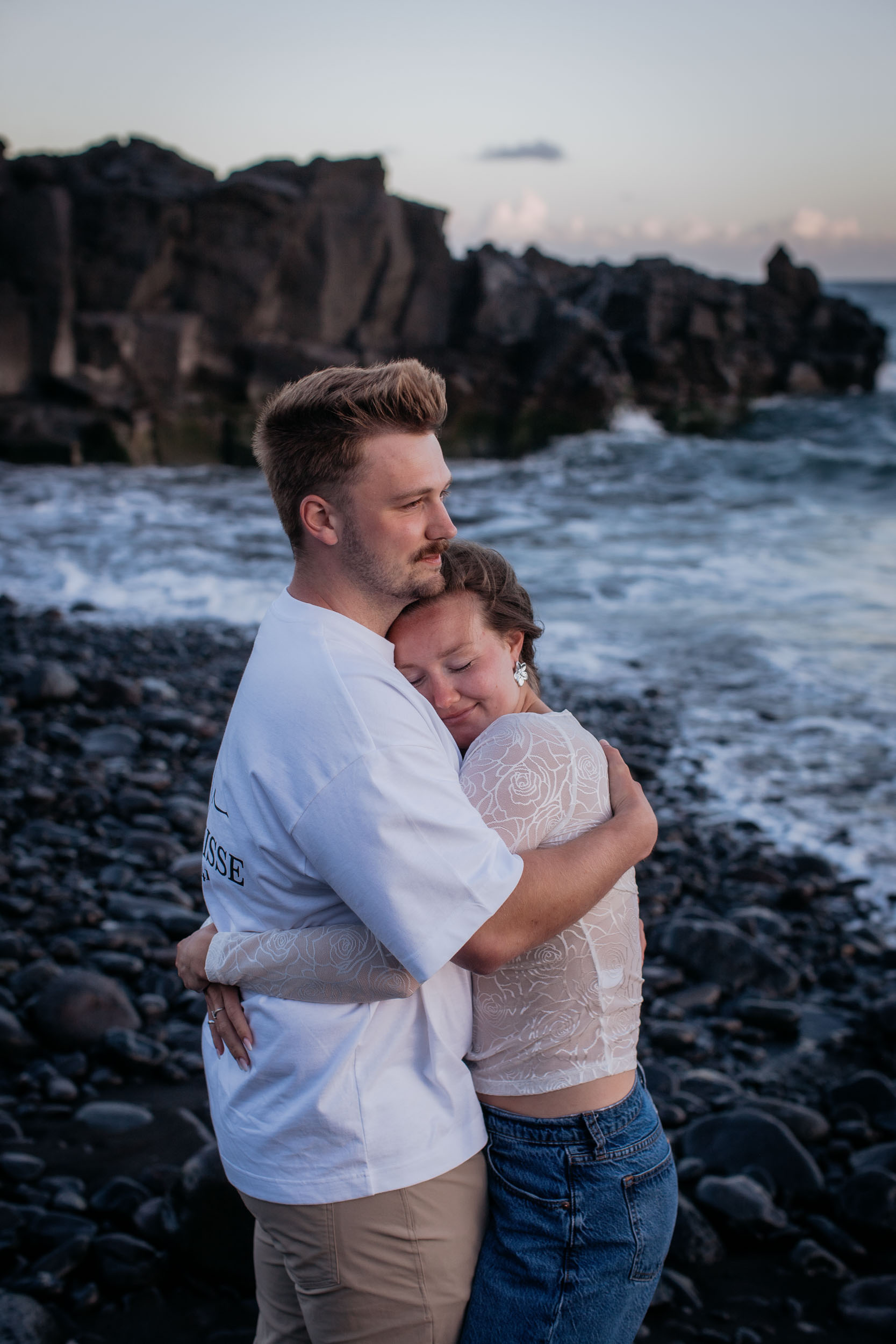 Rylie holding Abby close with her head on his chest and eyes closed, volcanic cliffs and white surf behind, Praia Formosa, Funchal, Madeira