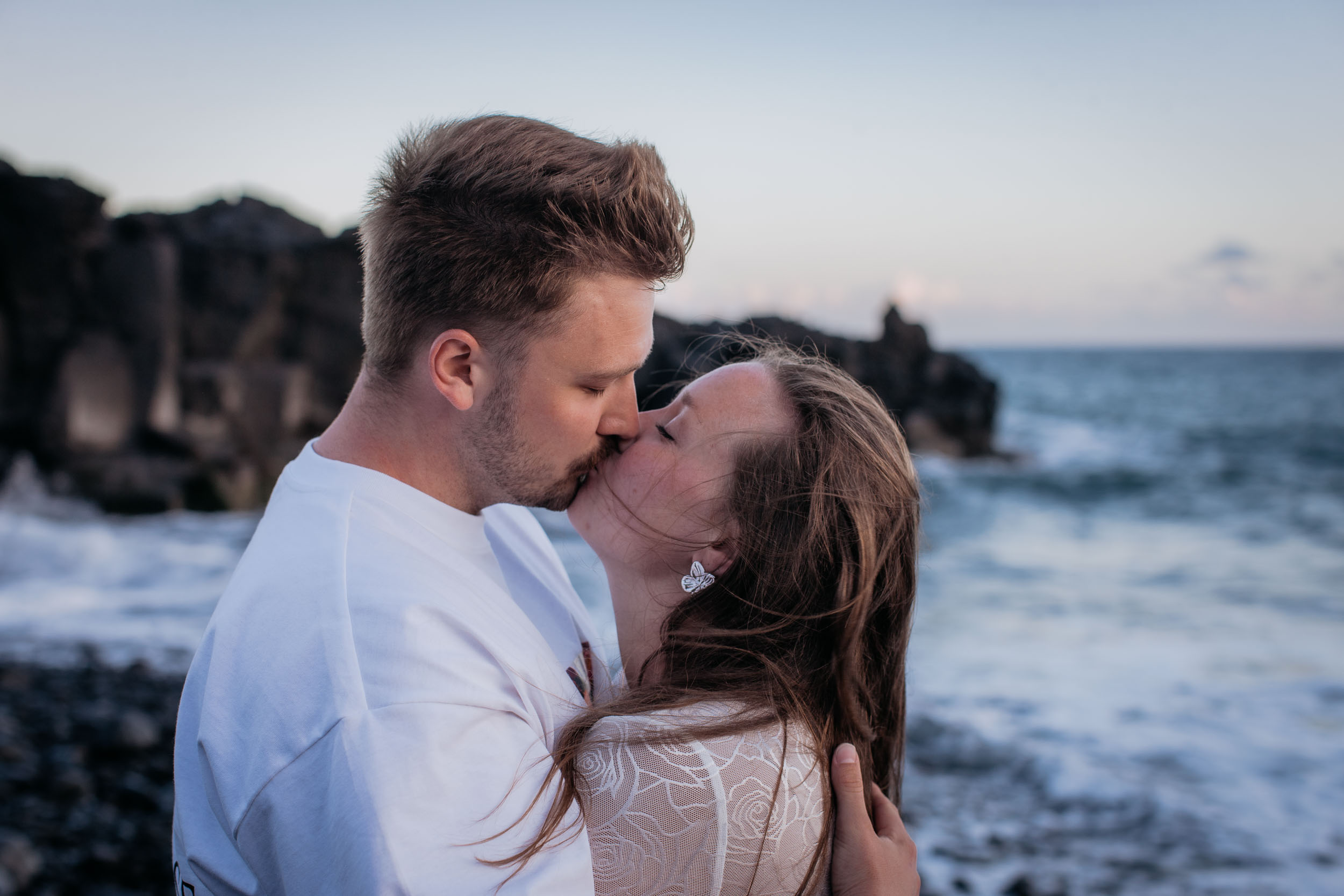Close portrait of Rylie kissing Abby on the forehead, volcanic cliffs and Atlantic water behind them at sunset, Praia Formosa, Madeira