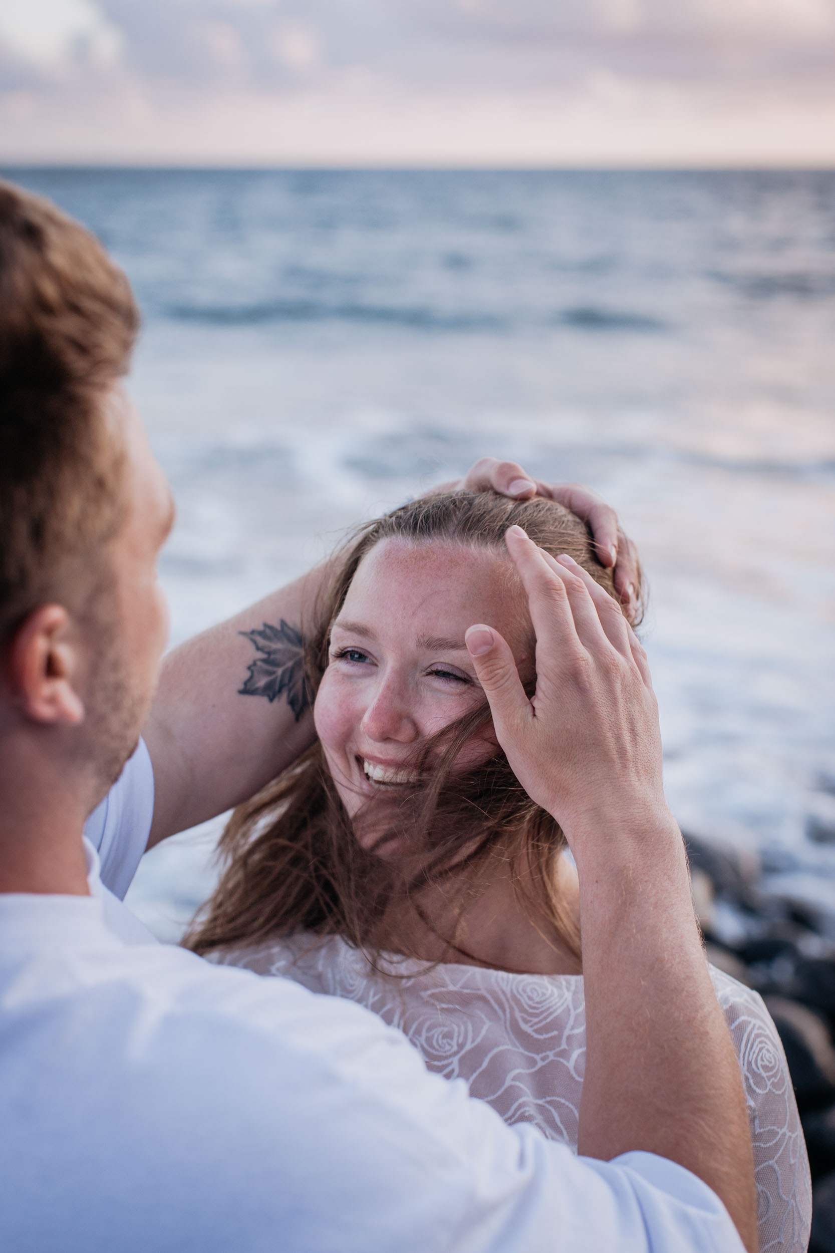 Vertical frame over his shoulder. He gathers her hair back with both hands and she laughs into the wind, Praia Formosa, Madeira