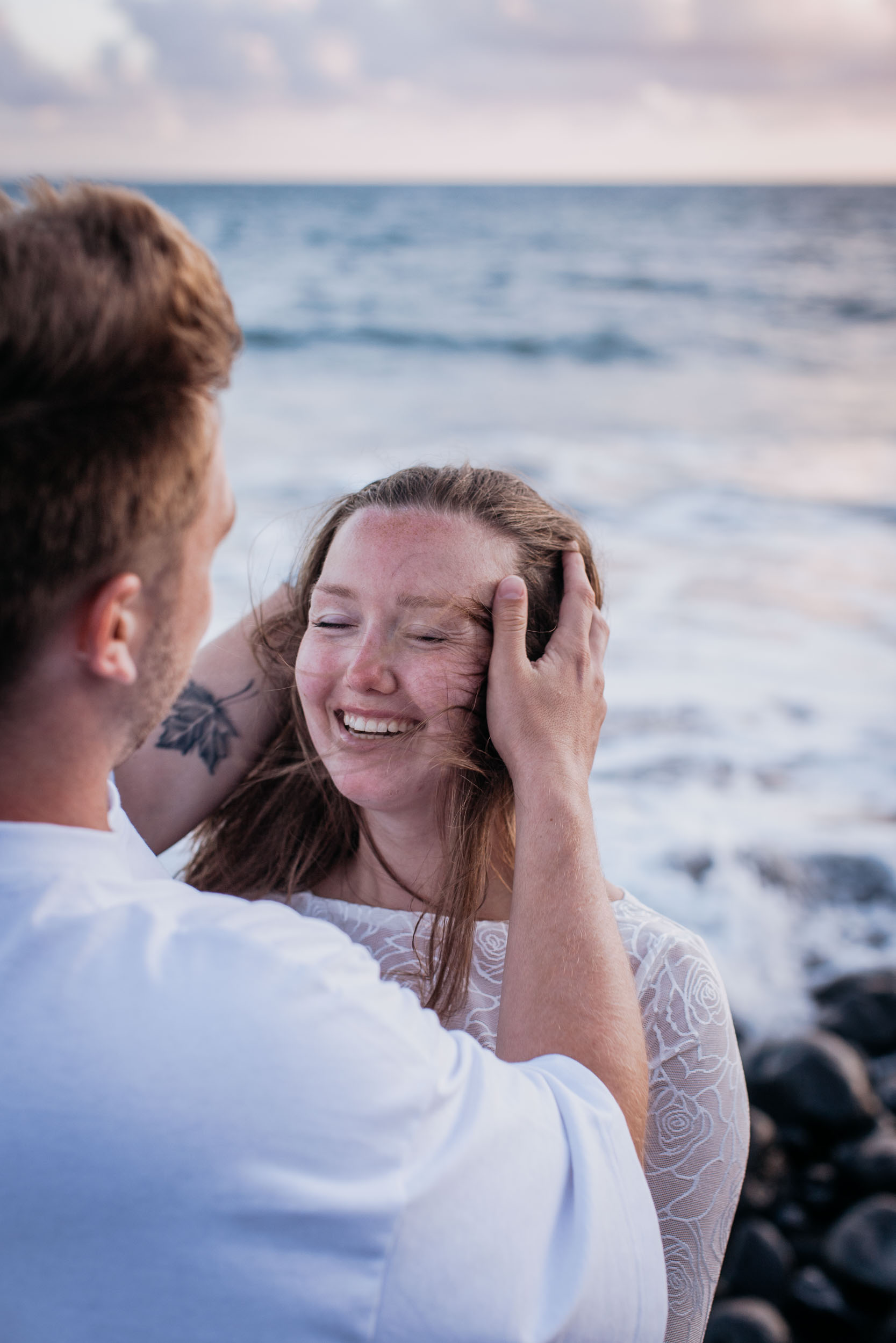 Vertical frame. He holds her face and she laughs with her eyes shut, his tattooed arm across the frame, Praia Formosa, Madeira