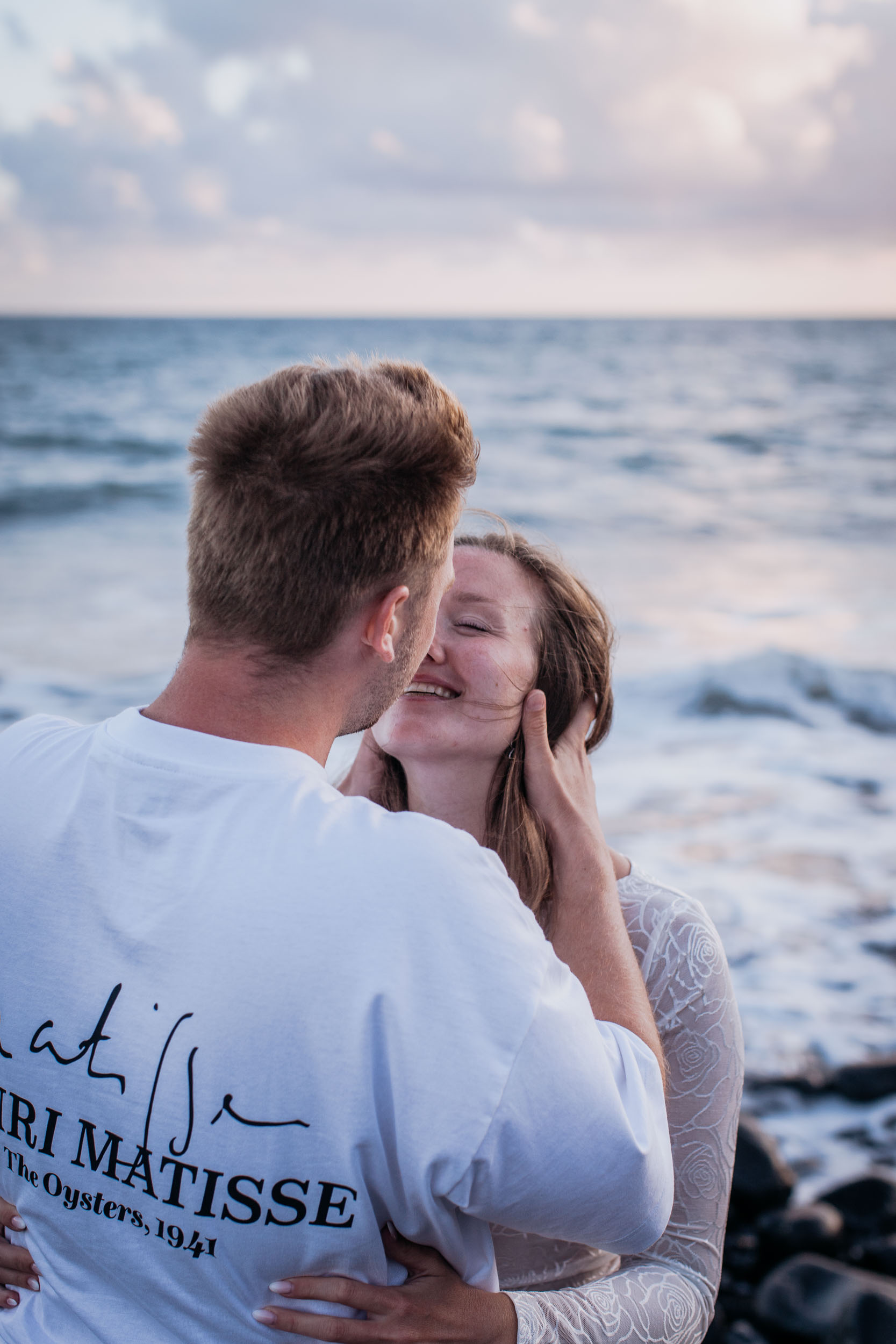 Vertical frame from behind his shoulder. She closes her eyes and smiles as he leans in, his hand at her cheek, Praia Formosa, Madeira
