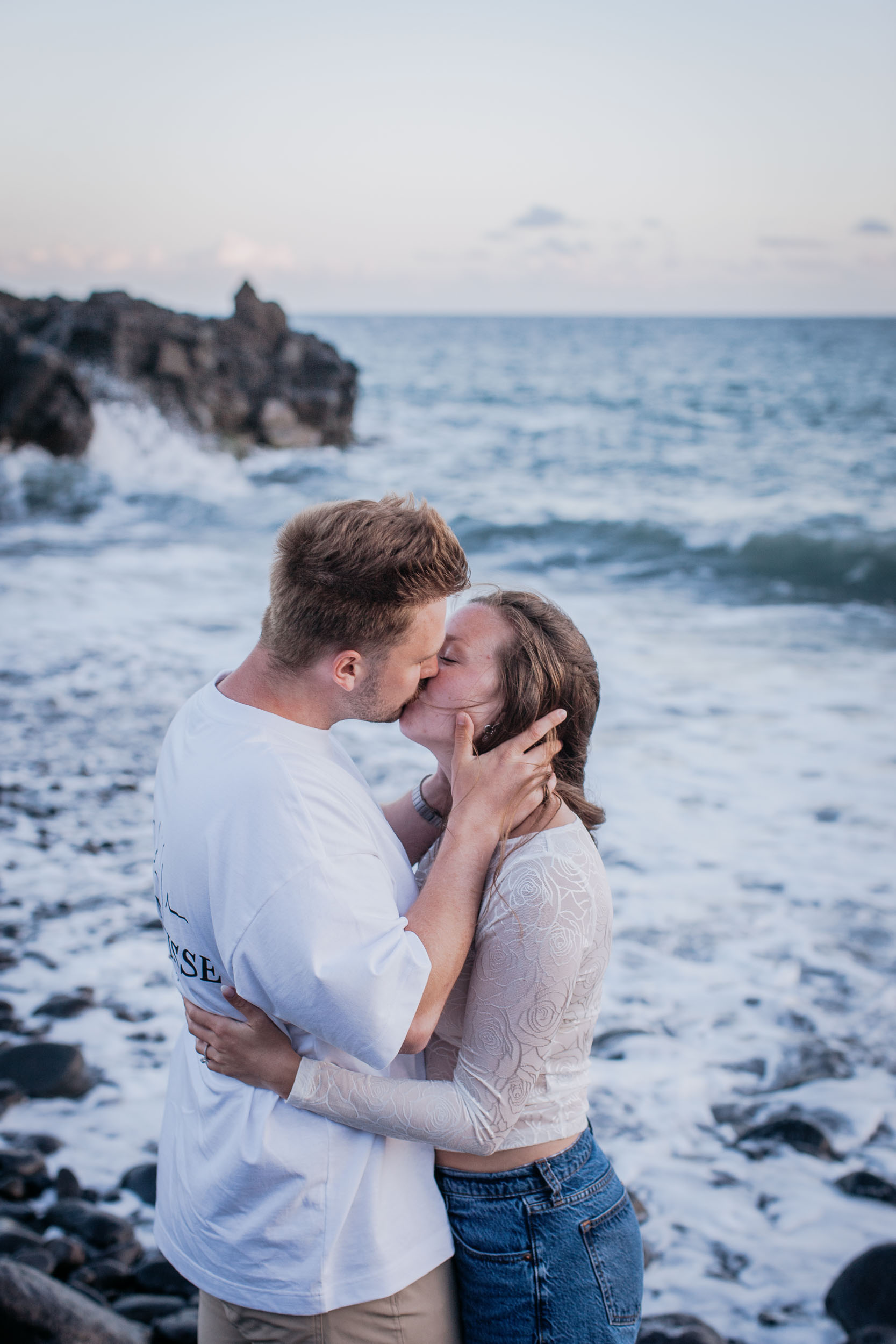 Vertical frame. A kiss on the stones, his hand at her jaw, a black sea stack and breaking surf behind, Praia Formosa, Madeira