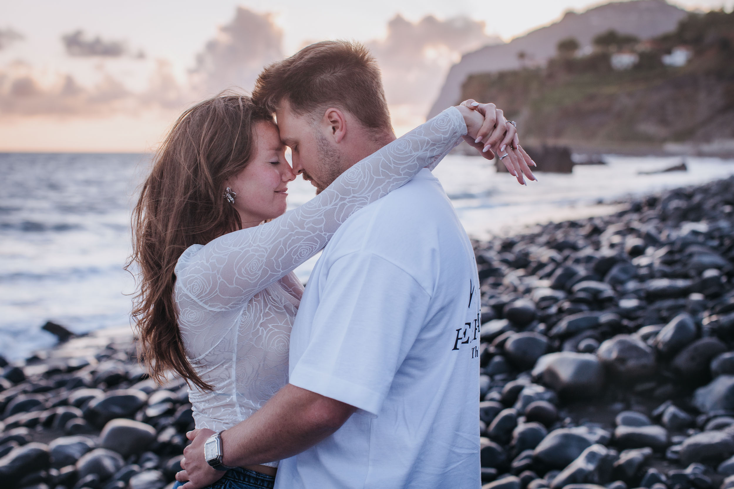 Rylie and Abby forehead to forehead with eyes closed, embracing on the pebble shore at Praia Formosa, Funchal cliffs behind, Madeira