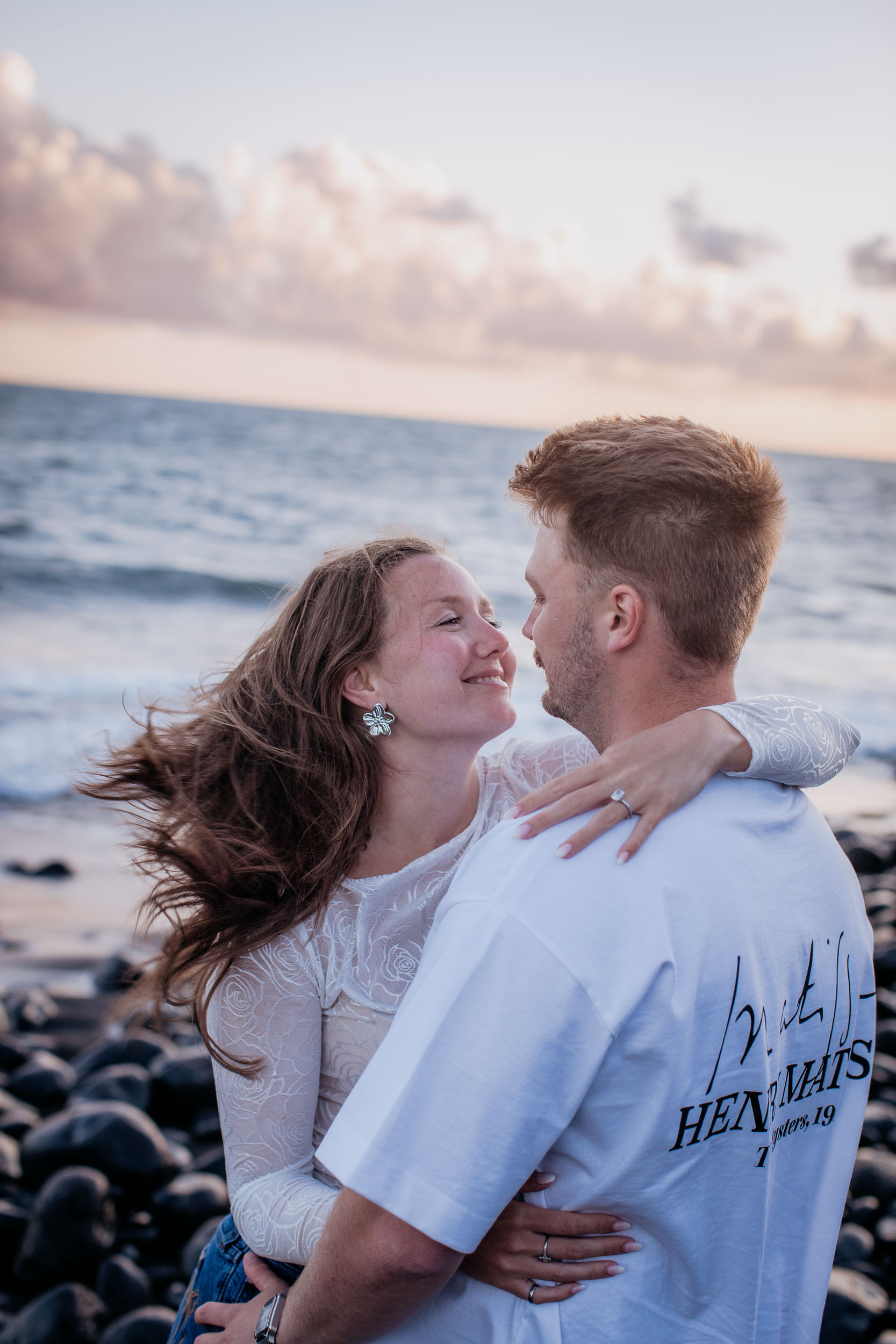 Vertical frame. She looks up at him smiling with her arm over his shoulder, ring on her hand, wind in her hair, Praia Formosa, Madeira