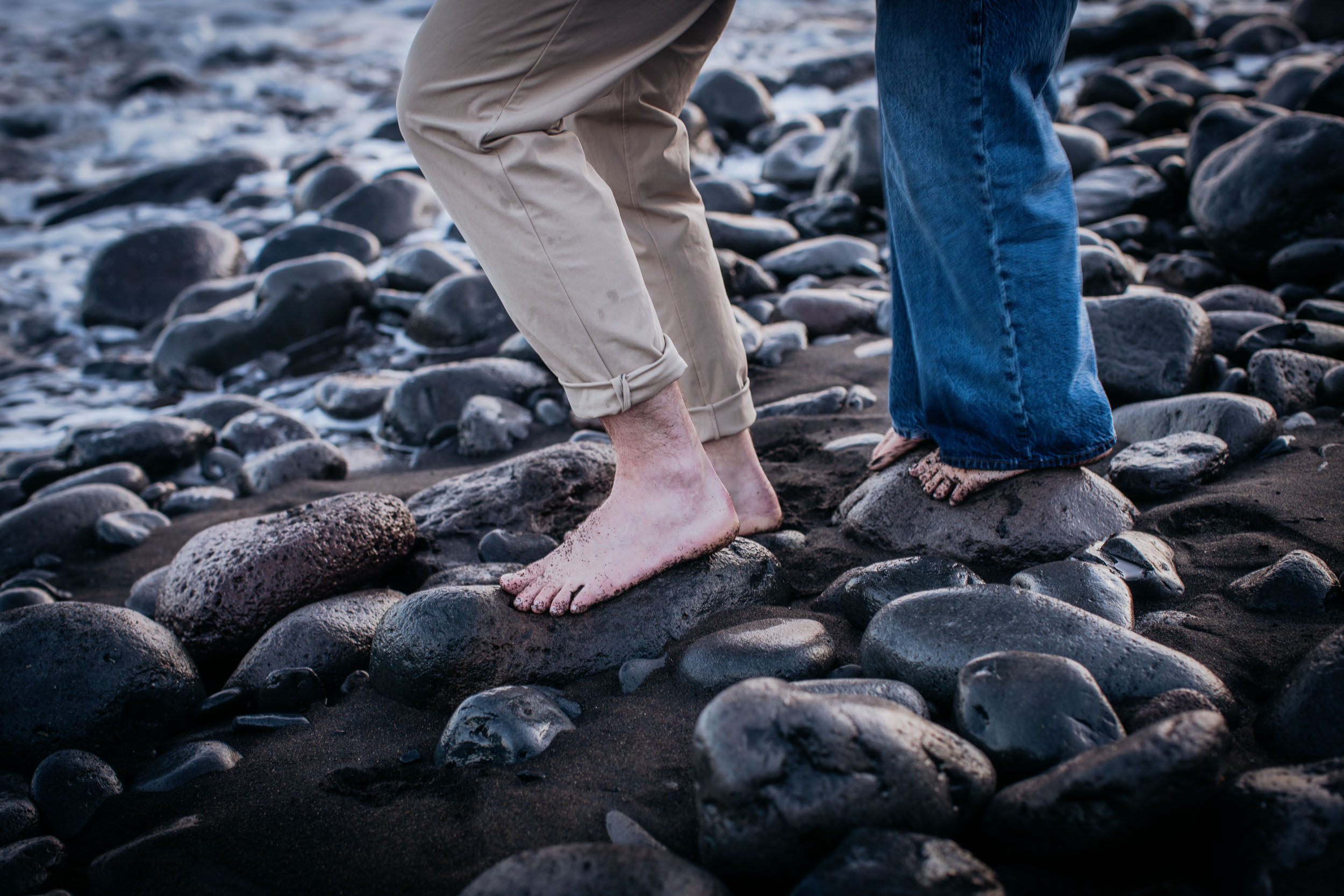 Detail. Four bare feet on wet volcanic pebbles, his trousers rolled at the ankle, Praia Formosa, Madeira