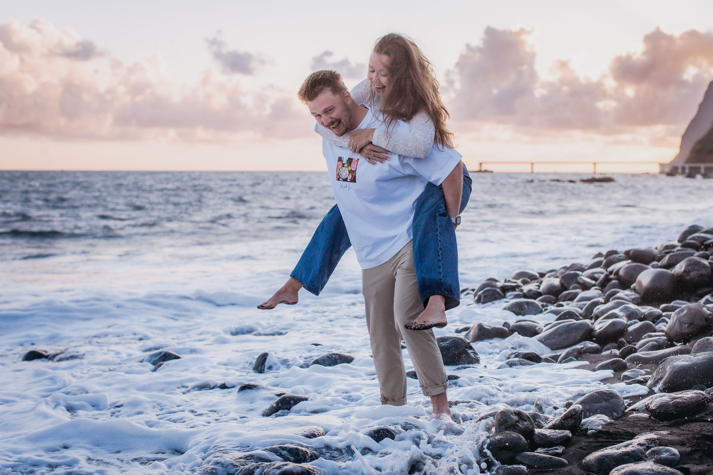 Rylie giving Abby a piggyback ride through shallow surf at Praia Formosa, both laughing, sunset light on the water, Funchal, Madeira