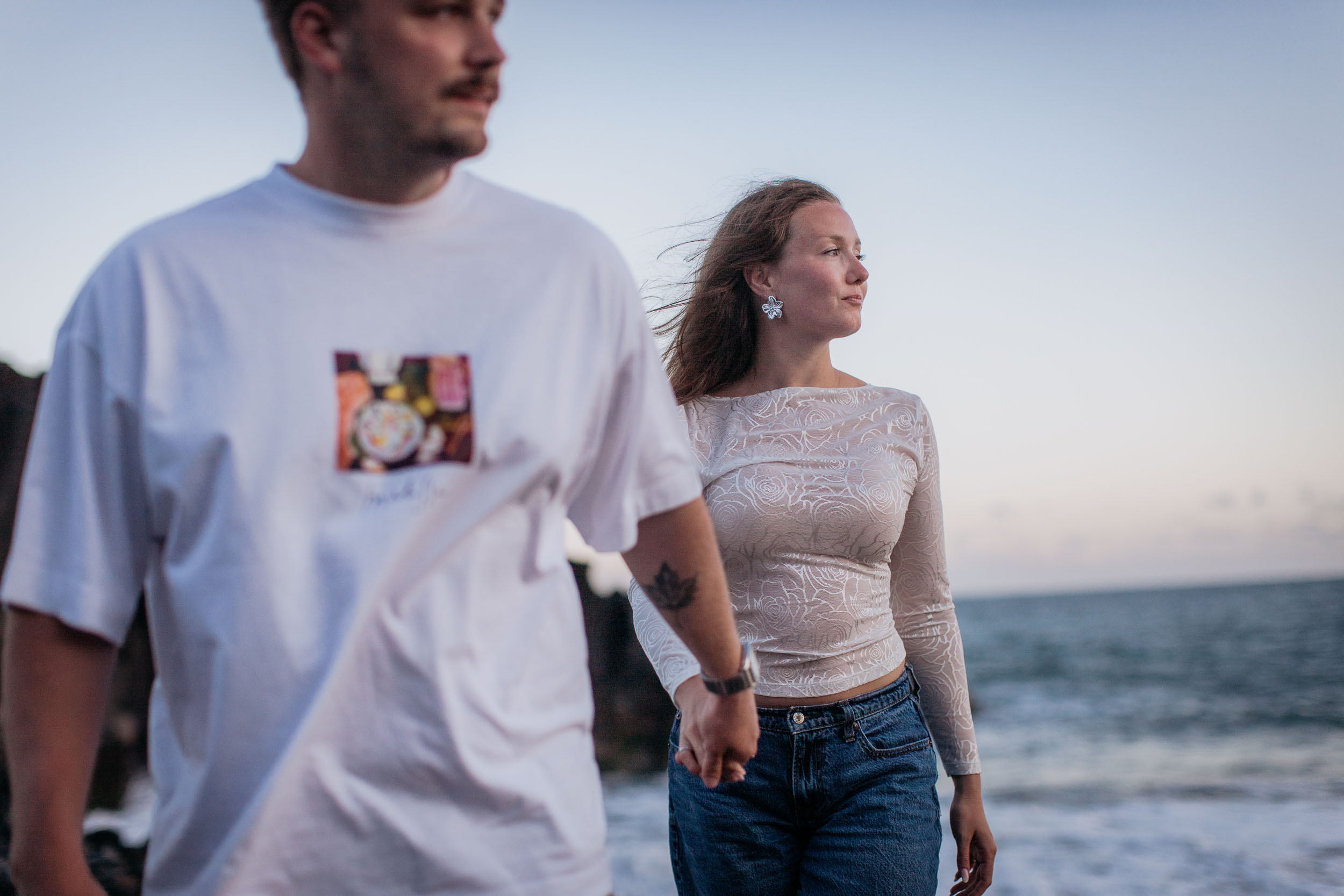 He passes close to the camera out of focus while she stays sharp behind, looking out to sea, hands joined, Praia Formosa, Madeira
