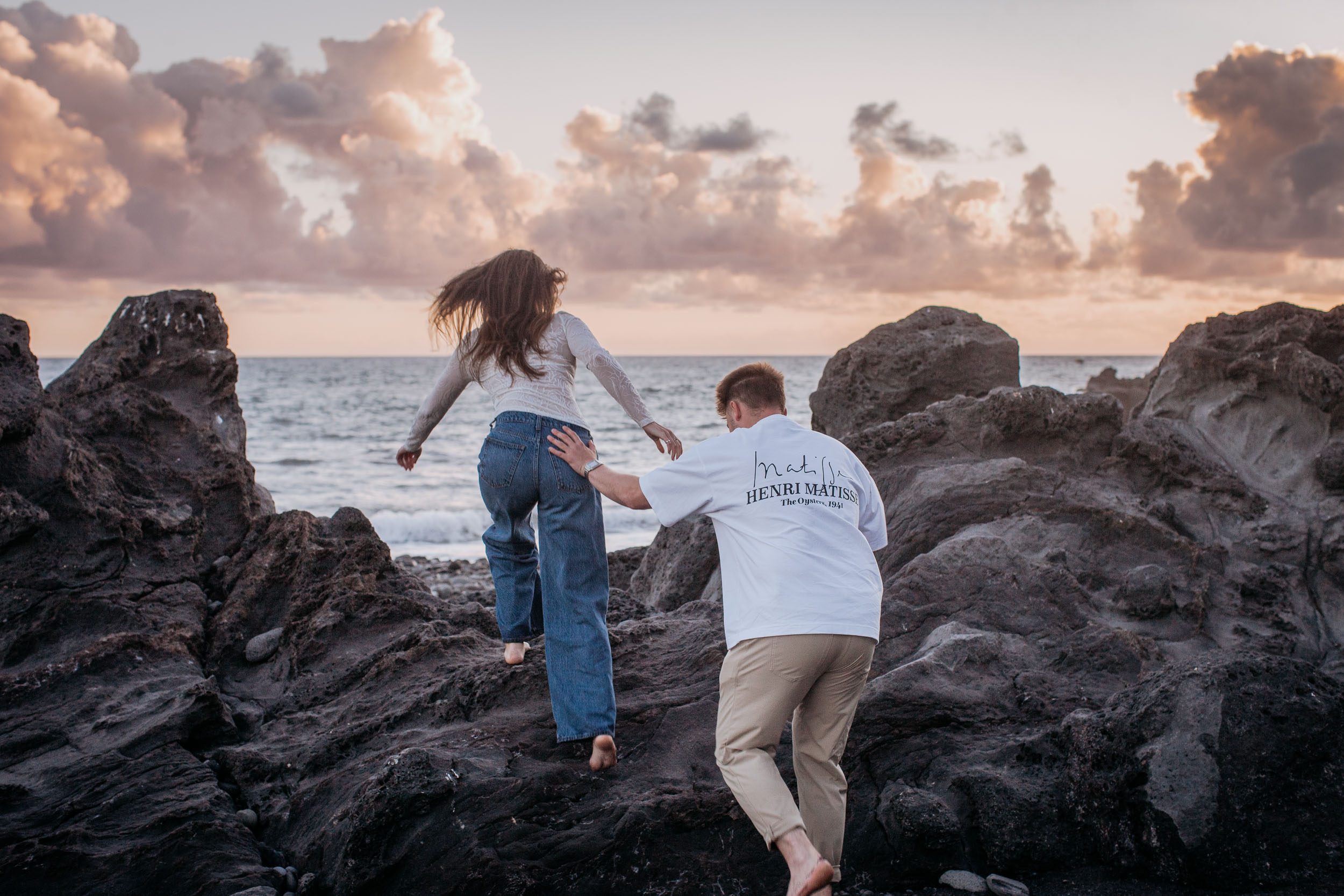 Seen from behind, he steadies her by the waist as she climbs the black lava rocks toward the water, Praia Formosa, Madeira