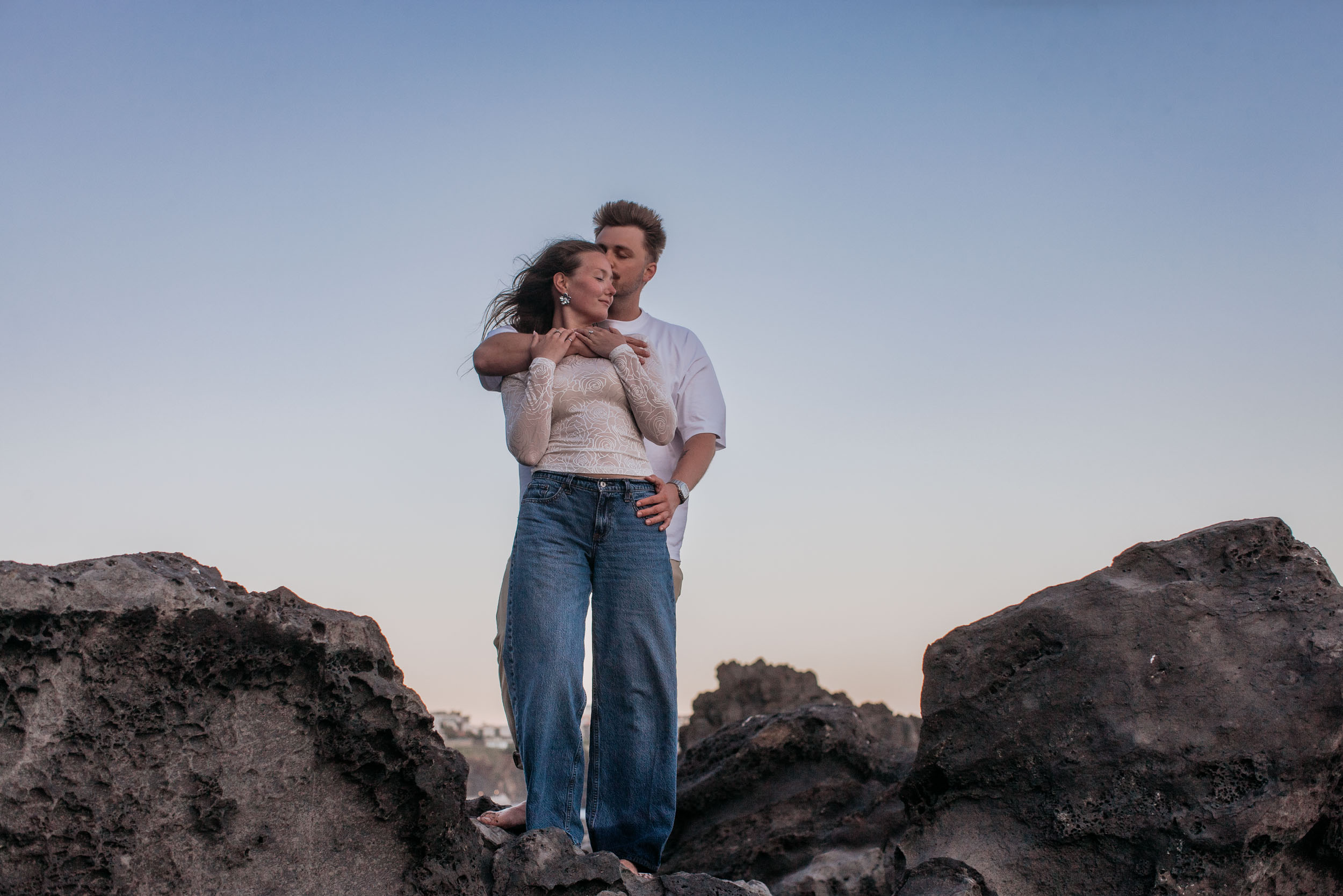 Rylie kissing the top of Abby's head as they stand together on a volcanic rock against an open dusk sky, Praia Formosa, Funchal, Madeira