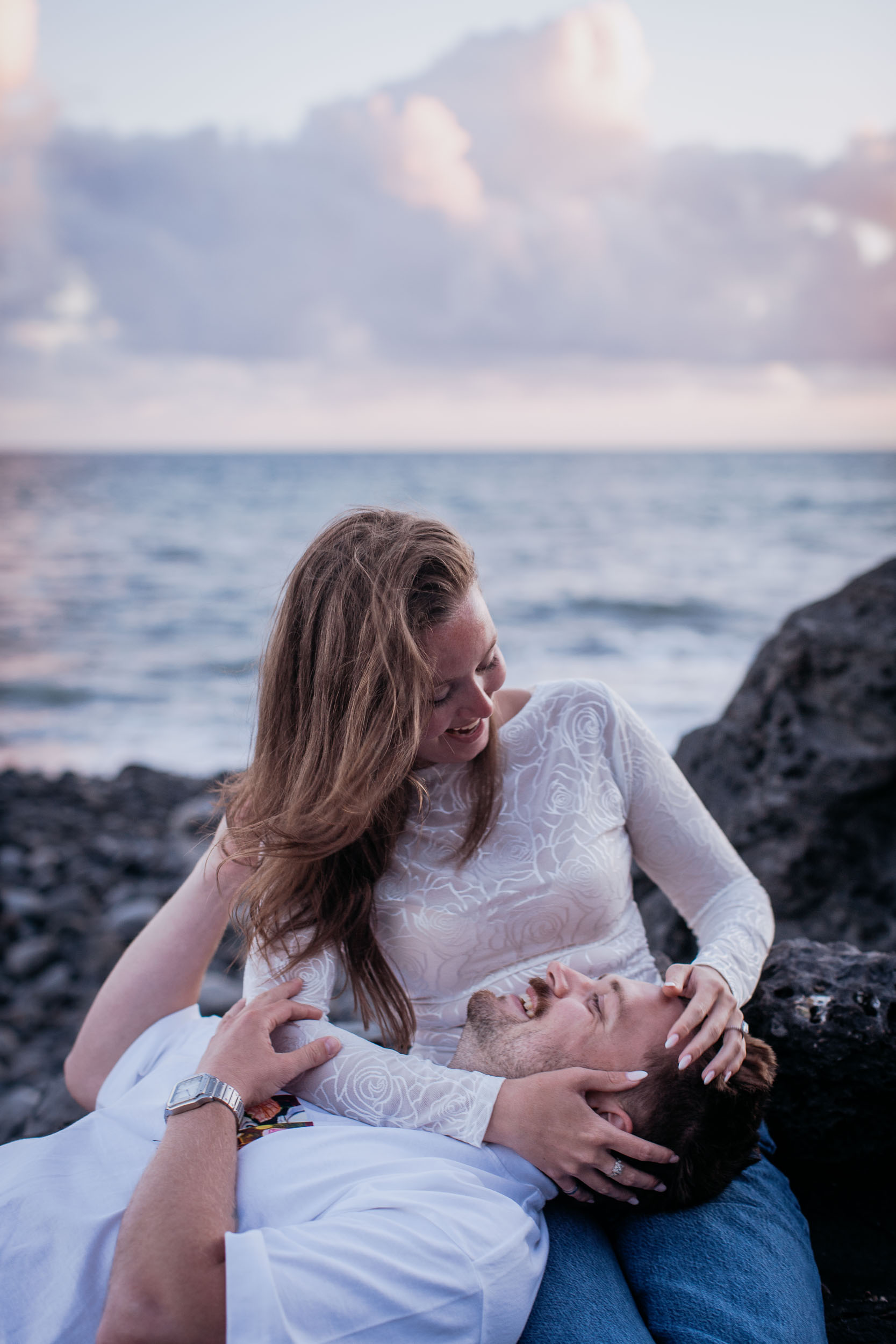 Vertical frame. He lies with his head in her lap on the rock, she looks down at him and holds his face, Praia Formosa, Madeira