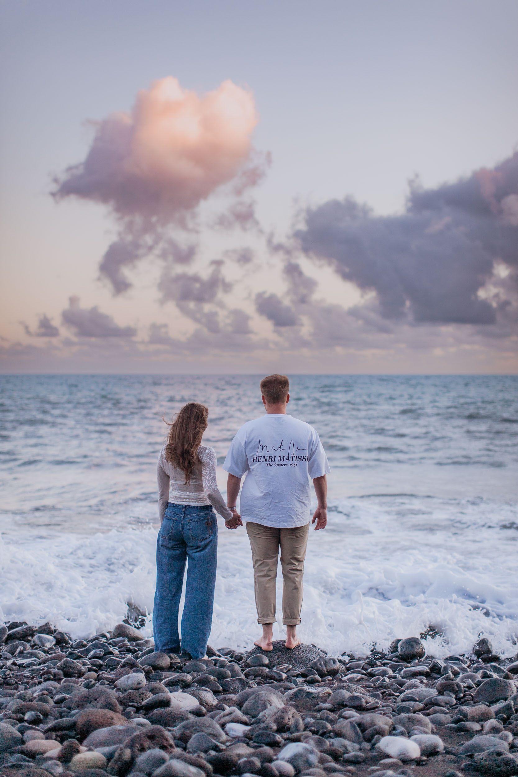 Rylie and Abby holding hands as they walk toward the ocean, a heart-shaped pink cloud above them in the dusk sky, Praia Formosa, Madeira