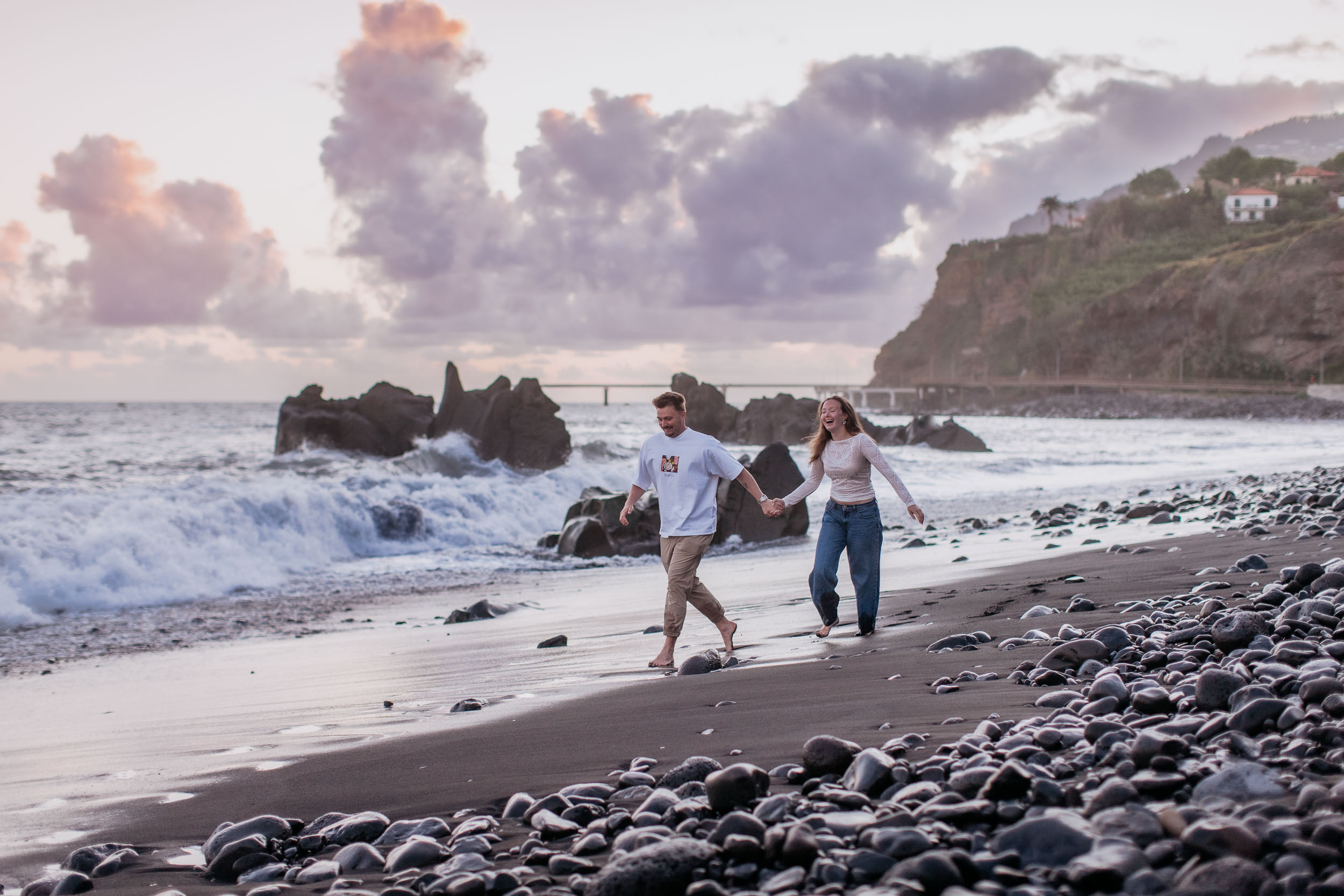 Rylie and Abby walking hand in hand along the black sand of Praia Formosa, breaking waves and the Funchal cliffs behind them at sunset, Madeira
