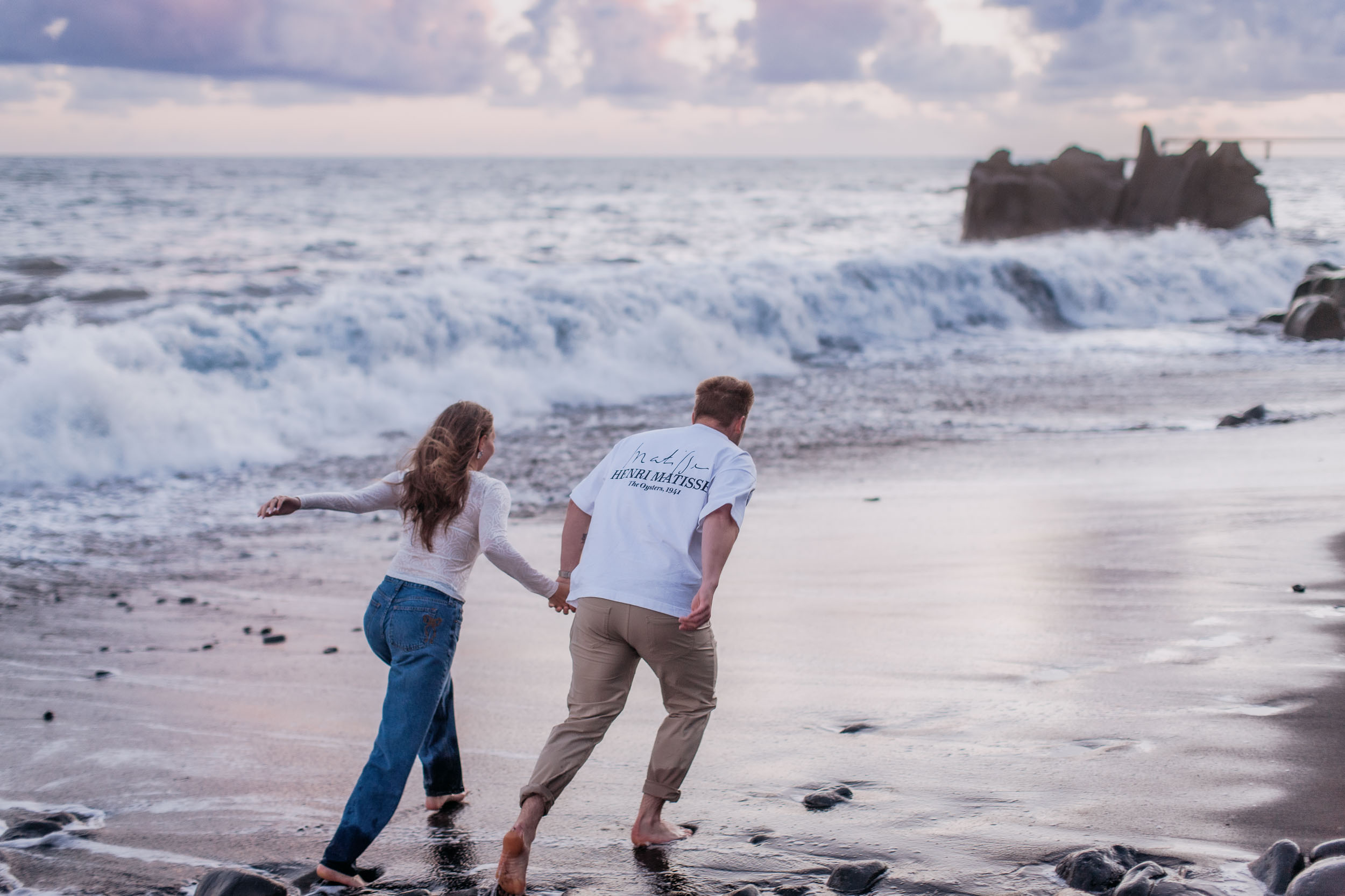 Rylie and Abby running hand in hand along wet black sand as a large wave breaks over the volcanic rocks behind them, Praia Formosa, Madeira
