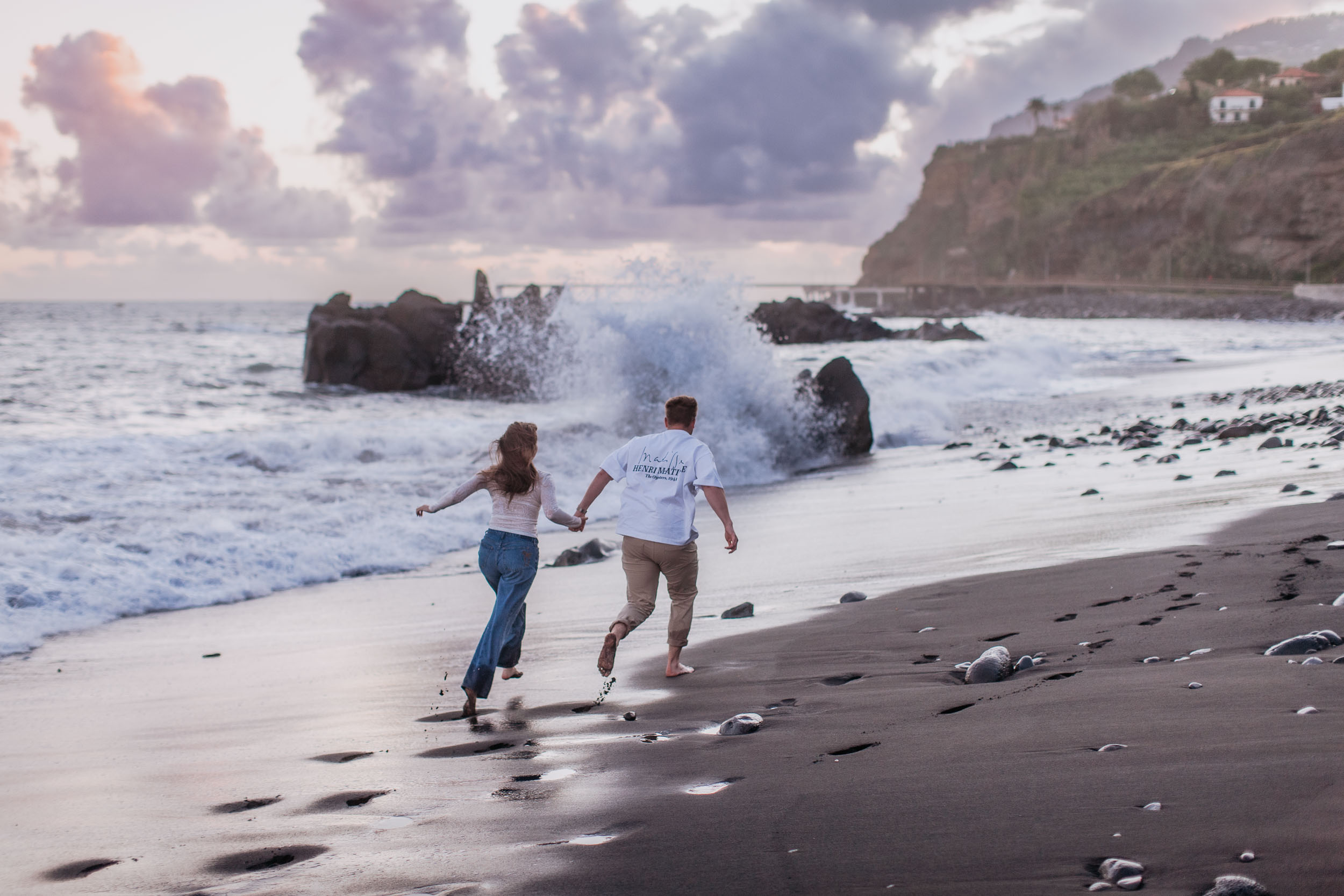 Rylie and Abby running barefoot along Praia Formosa's black sand, splash from a breaking wave and Funchal cliffs in the distance, Madeira
