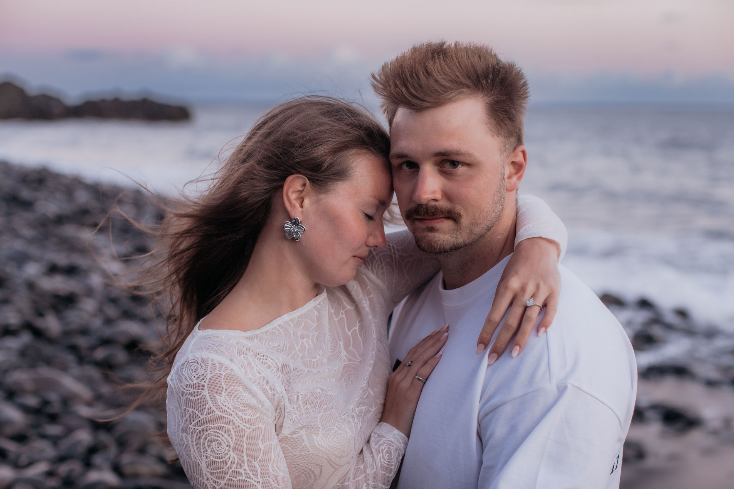 Close portrait of Rylie and Abby with foreheads touching, her eyes closed and his looking straight at the camera, dusk pink sky behind, Praia Formosa, Madeira