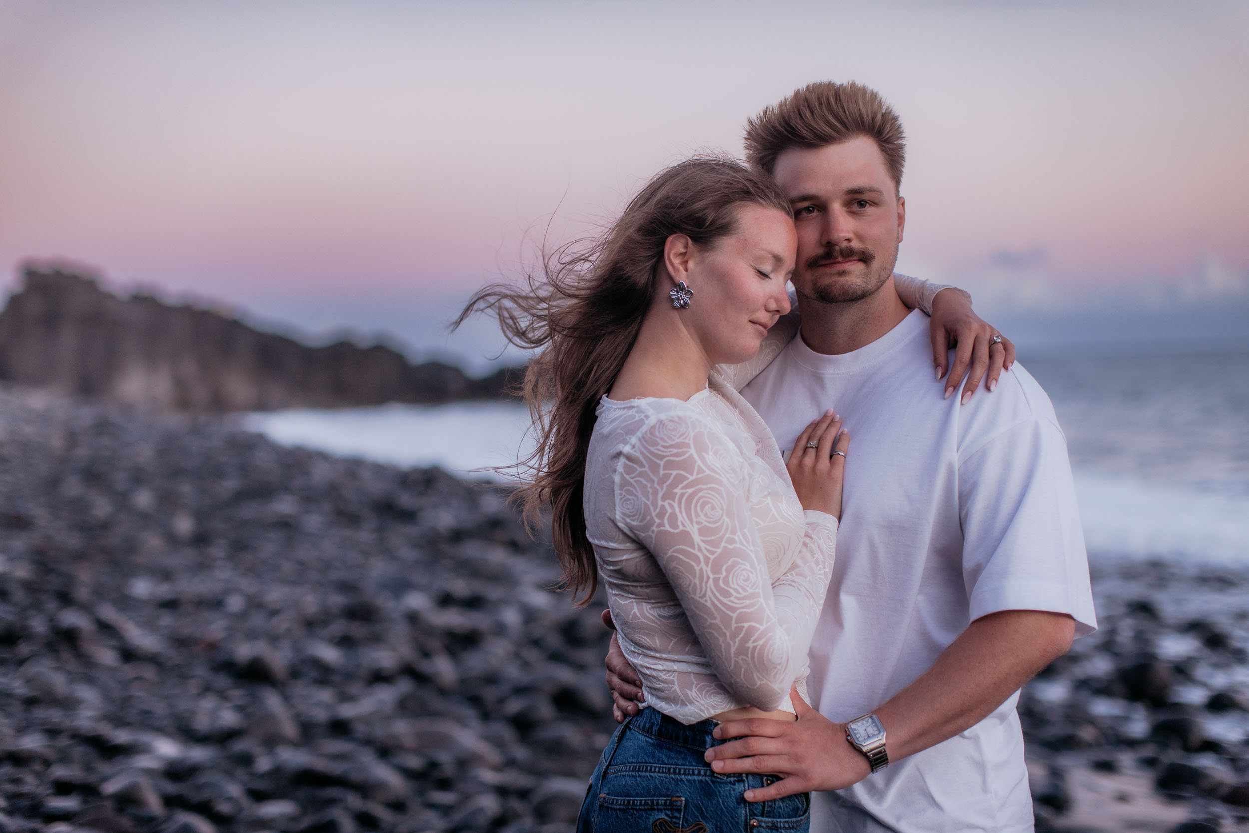 She rests her cheek on his shoulder with her eyes closed, he looks straight into the camera, pink dusk behind, Praia Formosa, Madeira