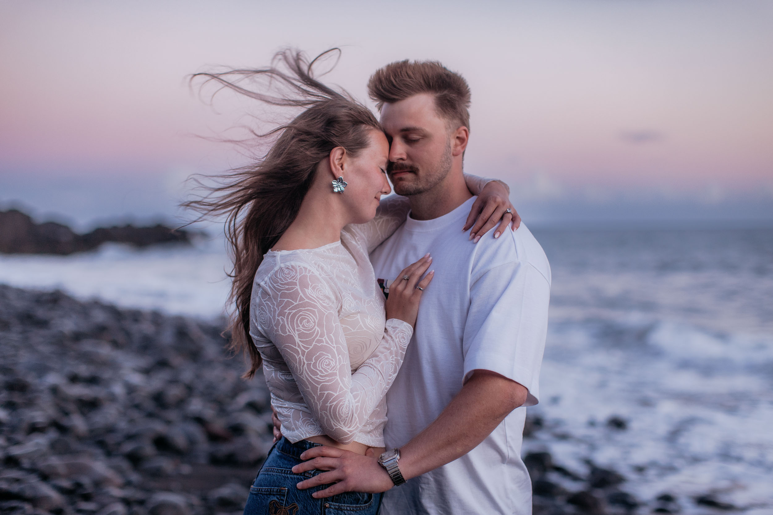 Forehead to forehead with eyes closed, her hand flat on his chest, wind pulling her hair sideways, Praia Formosa, Madeira