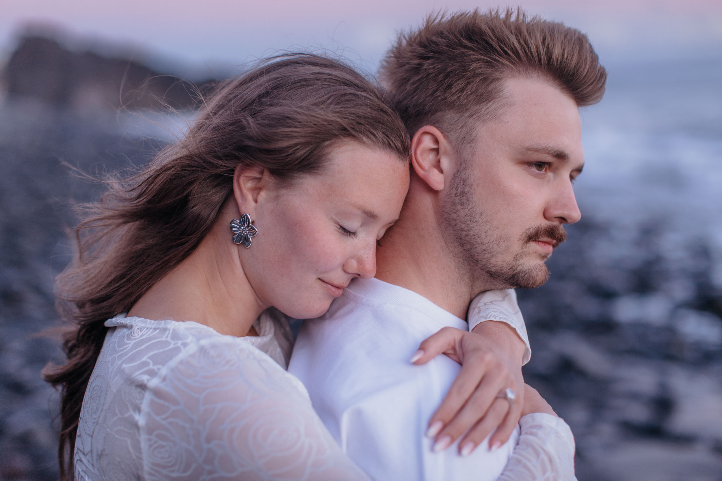 Abby leaning into Rylie's shoulder with eyes closed, his arm around her, dusk light and breaking surf behind, Praia Formosa, Madeira