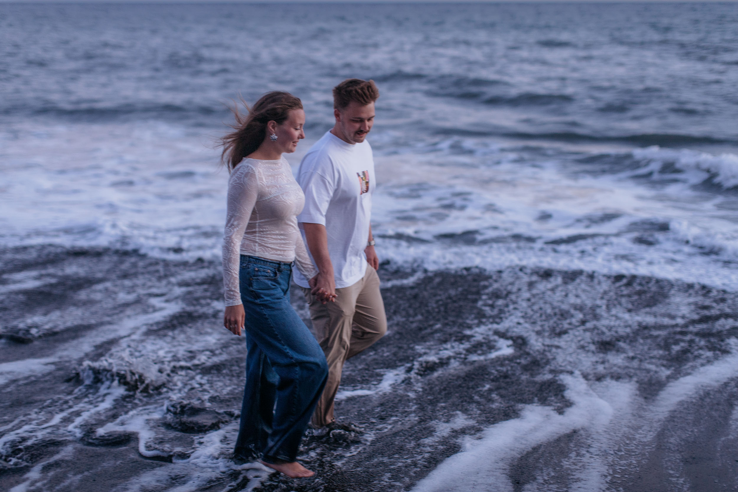 Walking hand in hand through the shallow foam, both looking down at the water, Praia Formosa, Madeira