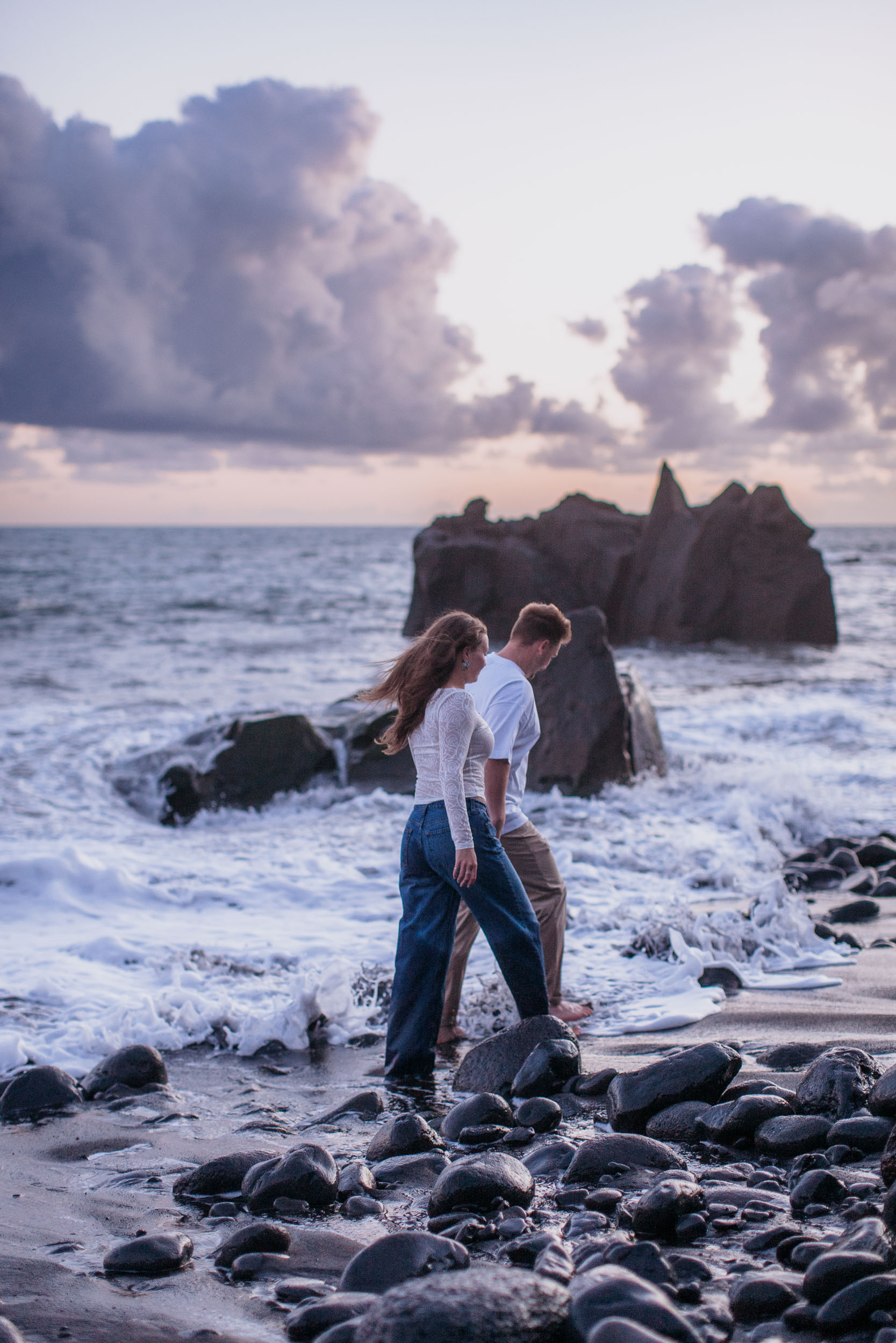 Rylie and Abby wading through shallow water by a tall volcanic rock formation, dramatic dusk clouds above, Praia Formosa, Madeira