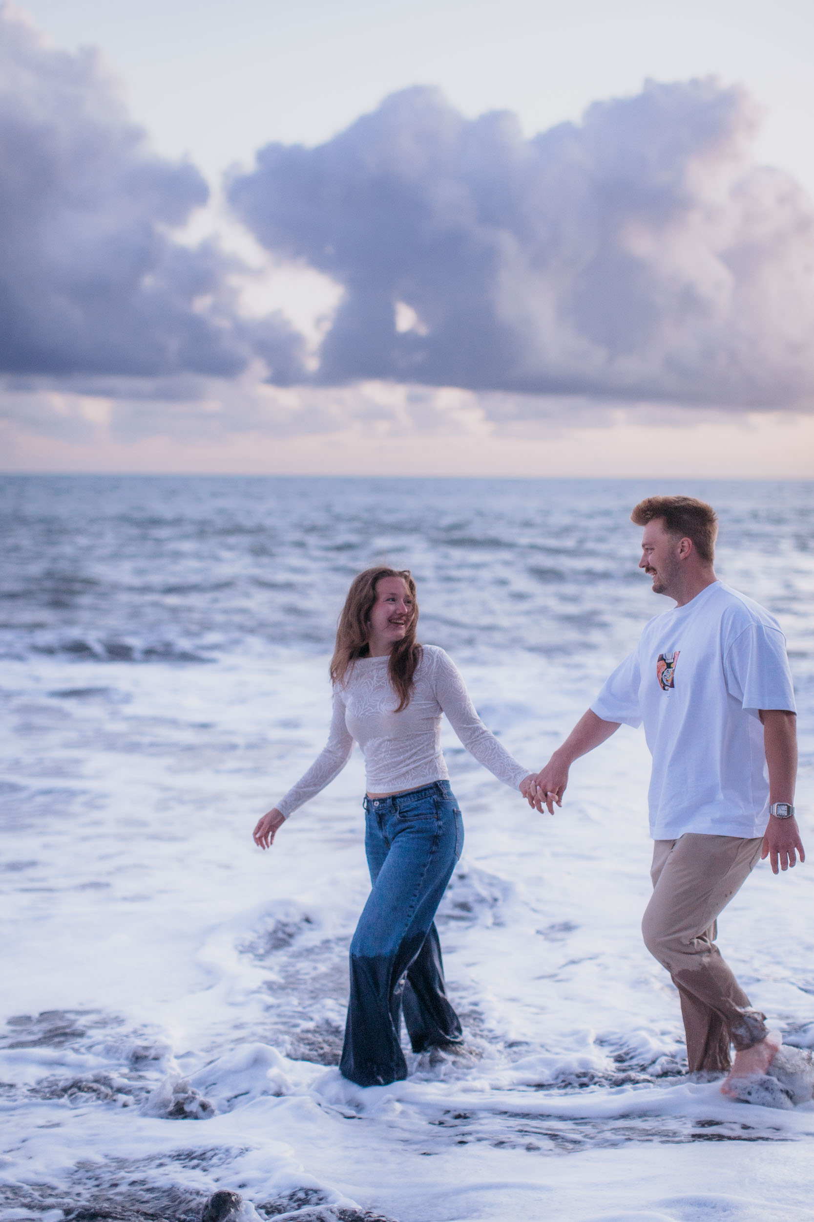 Rylie pulling Abby by the hand as they wade laughing into the white surf at Praia Formosa, evening clouds behind, Funchal, Madeira
