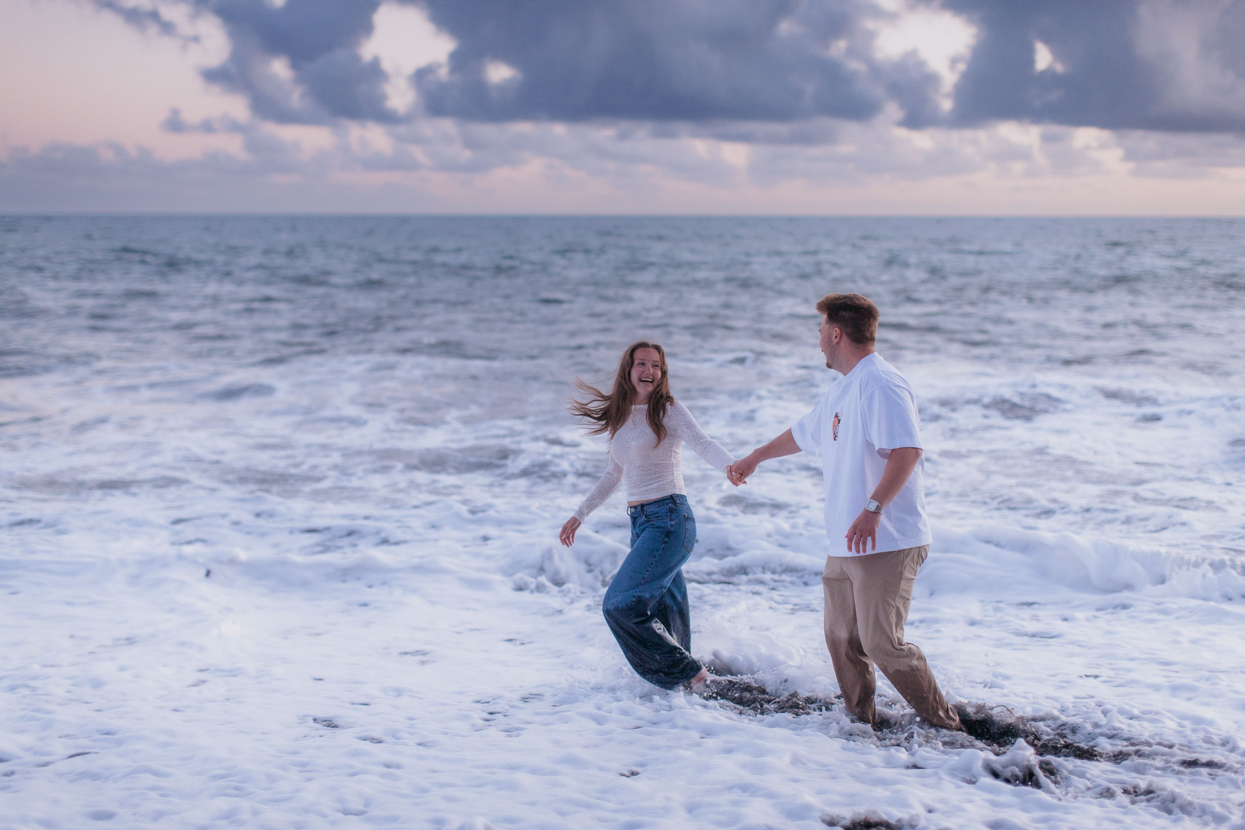 Couple laughing together while sea foam surges around their legs at Praia Formosa, dusk light, Madeira